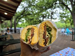 Cross-section view of a sandwich with sausage and greens, in front of a park-like setting with green trees