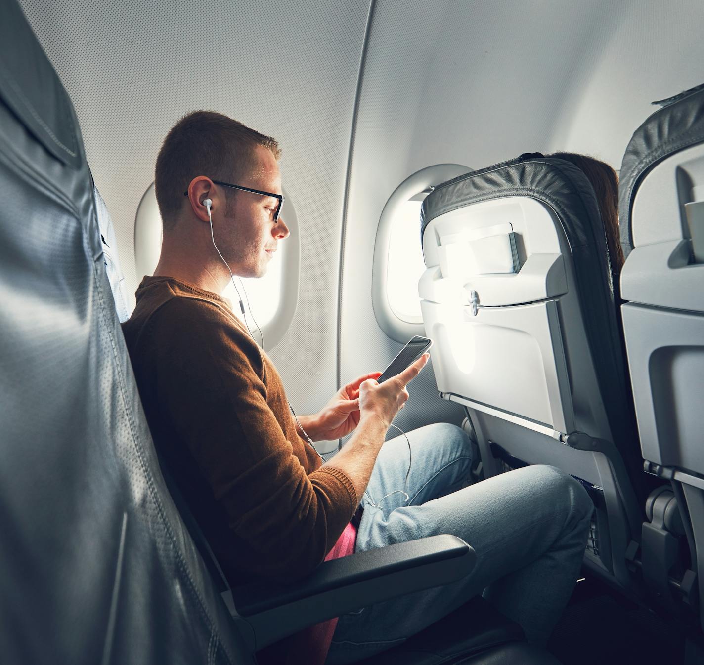 Connection in the airplane. Young man (traveler) using smart phone during flight and listening music.