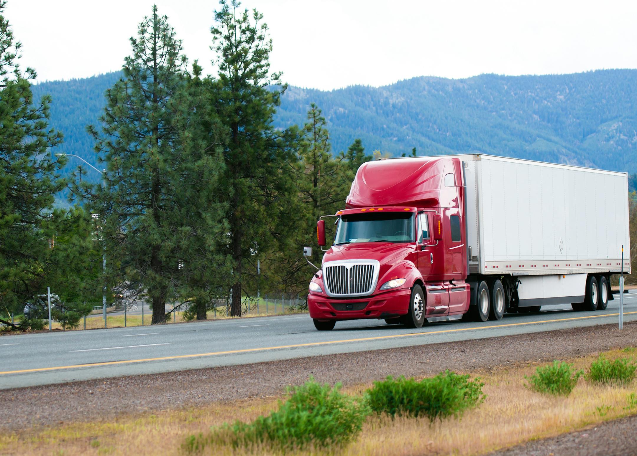 Red big rig modern shiny semi-truck with white dry van trailer with aerodynamic skirt move on straight divided interstate highway I-5 in California with green trees and mountains on background
