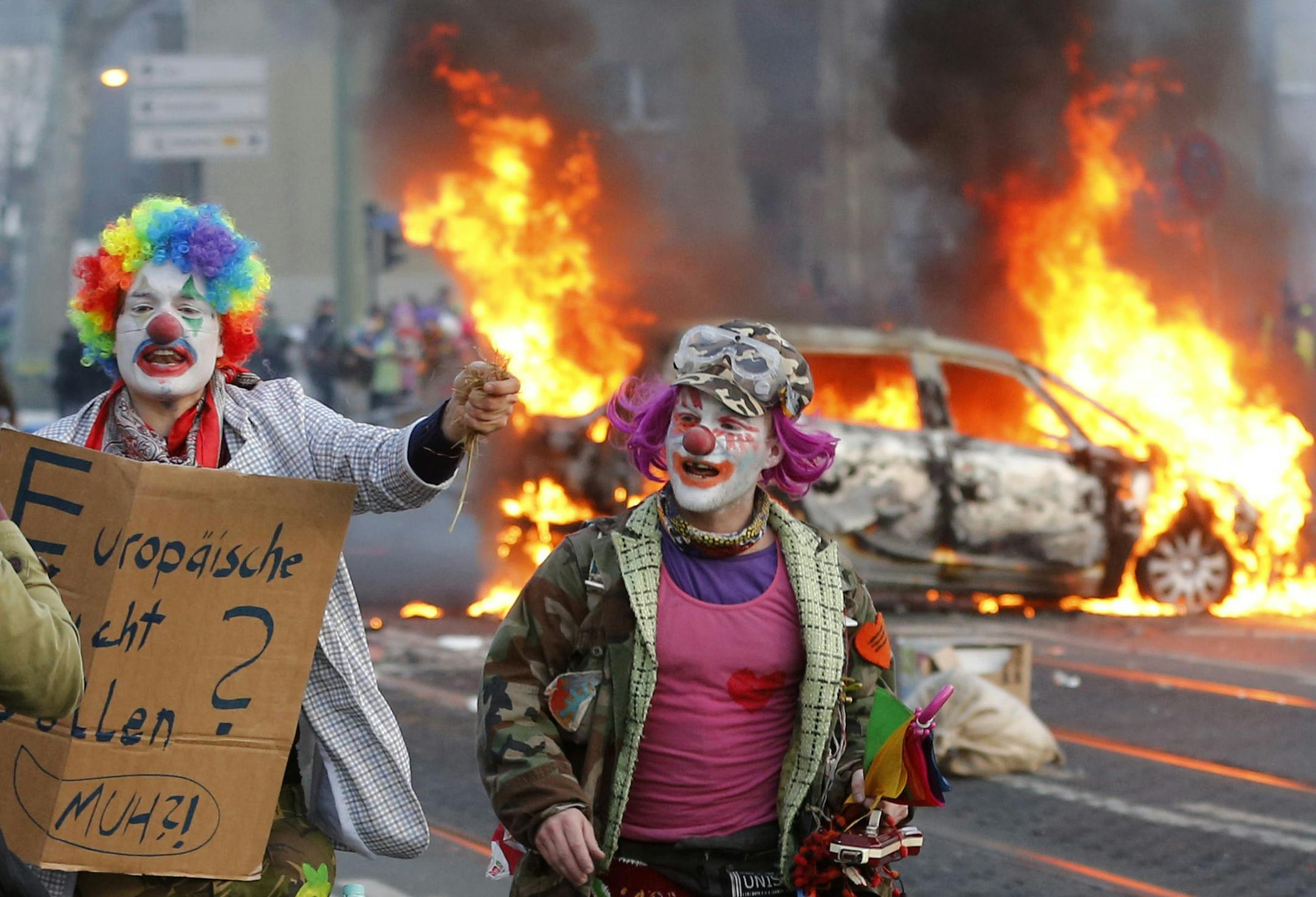 Demonstrators dressed as clowns pass by a burning police car Wednesday, March 18, 2015 in Frankfurt, Germany. Blockupy activists try to blockade the new headquarters of the ECB to protest against government austerity and capitalism. (AP Photo/Michael Probst)