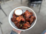 A white bowl of dry rub wings. The wings have a tanned, orangey hue with black specks of pepper and a char roasted jalepeno in the middle. On the side is a container of queso fresco ranch for dipping.