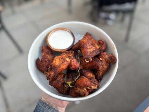 A white bowl of dry rub wings. The wings have a tanned, orangey hue with black specks of pepper and a char roasted jalepeno in the middle. On the side is a container of queso fresco ranch for dipping.