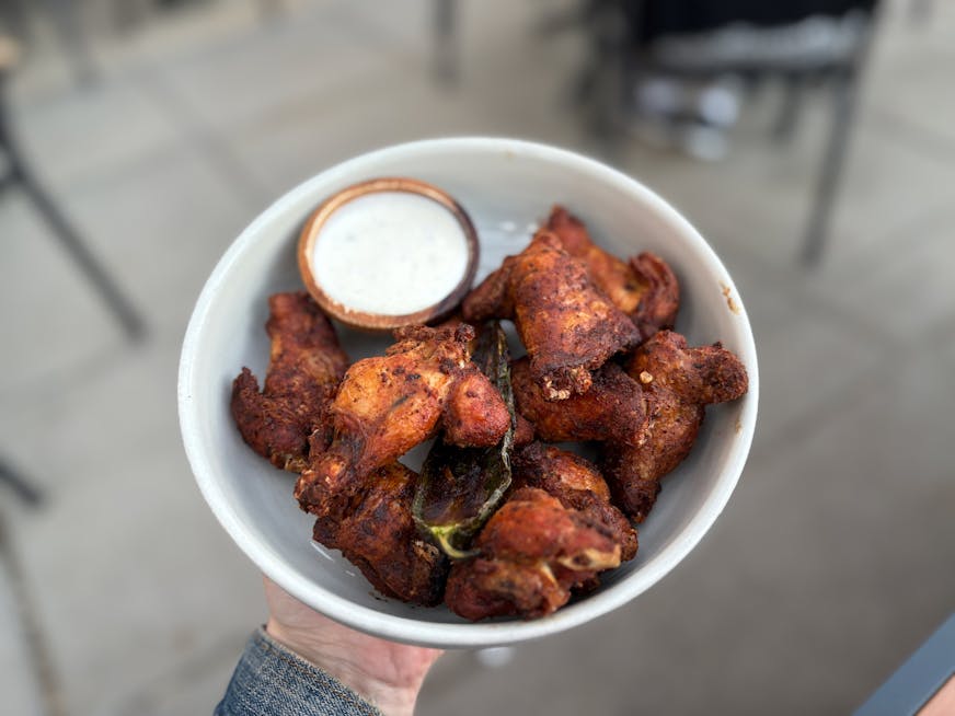 A white bowl of dry rub wings. The wings have a tanned, orangey hue with black specks of pepper and a char roasted jalepeno in the middle. On the side is a container of queso fresco ranch for dipping.