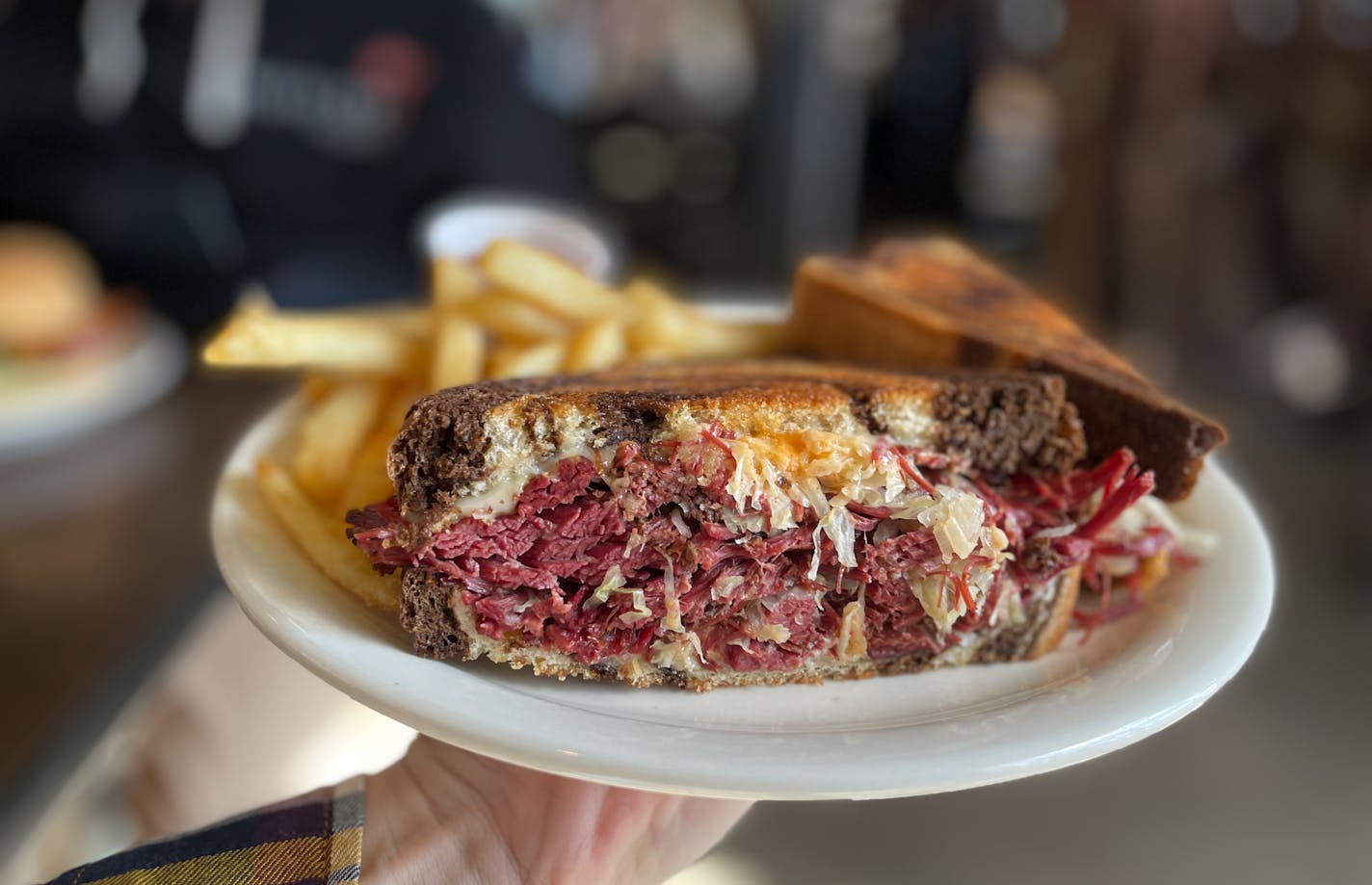 A hand holds a plate with a Rueben: marble rye bread, thick layers of corned beef with visible sauerkraut and thousand island dressing in the Urban Growler taproom.