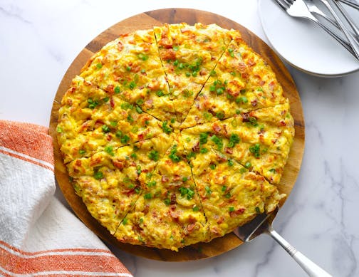 A round hash brown frittata with bacon and cheddar cheese on a wooden cutting board with a serving piece.