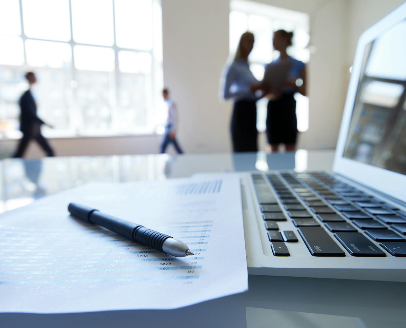 Document with statistical data, laptop and pen on desk, business people in the background