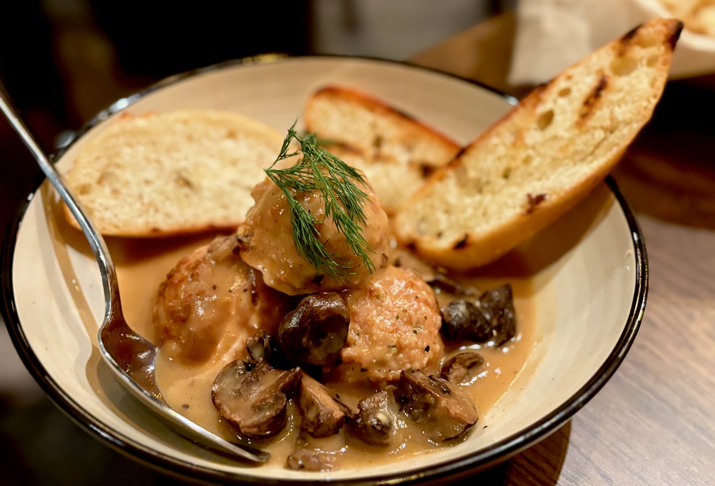 A shallow bowl with a stack of light colored meatballs garnished with a sprig of dill over mushrooms in gravy. Two slices of charred bread are tucked into the back of the bowl.