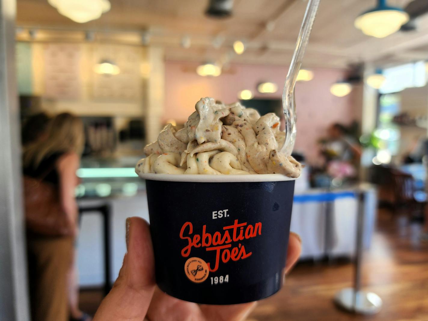 A cup that says "Sebastian Joe's" with soft vanilla ice cream flecked with rainbow colors, held up inside a busy shop