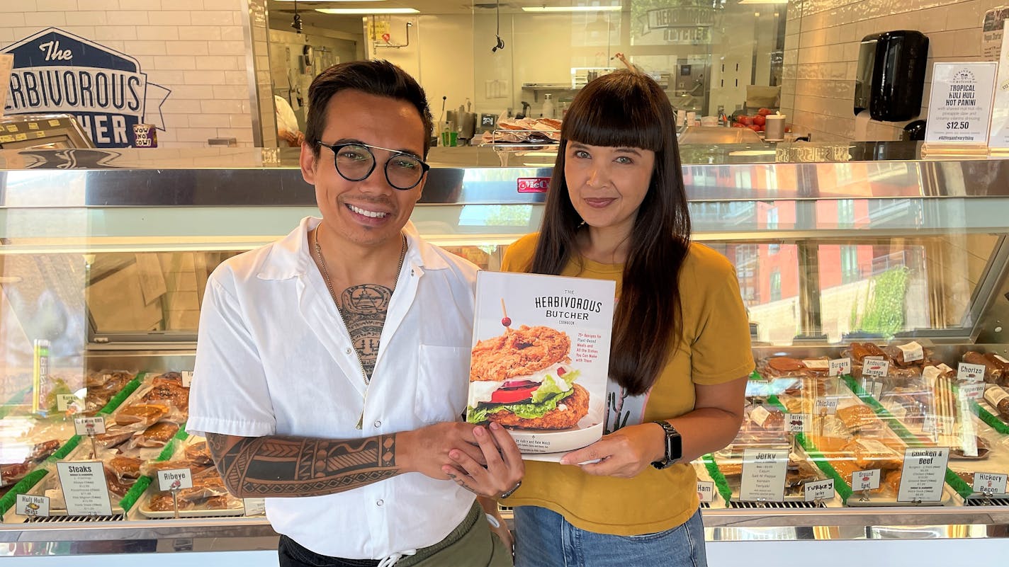 Kale Walch and Aubry Walch holding their cookbook in front of a case of vegan meat at their store