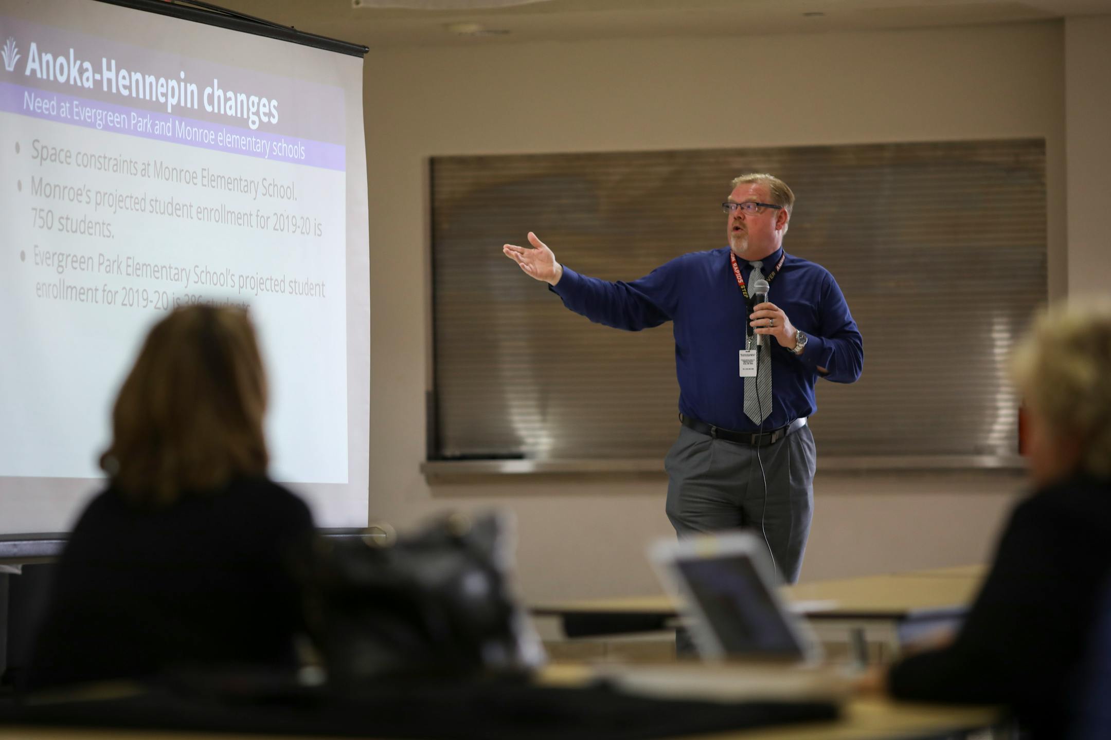 A man in a shirt and tie gestures to a slideshow projected on a screen behind him