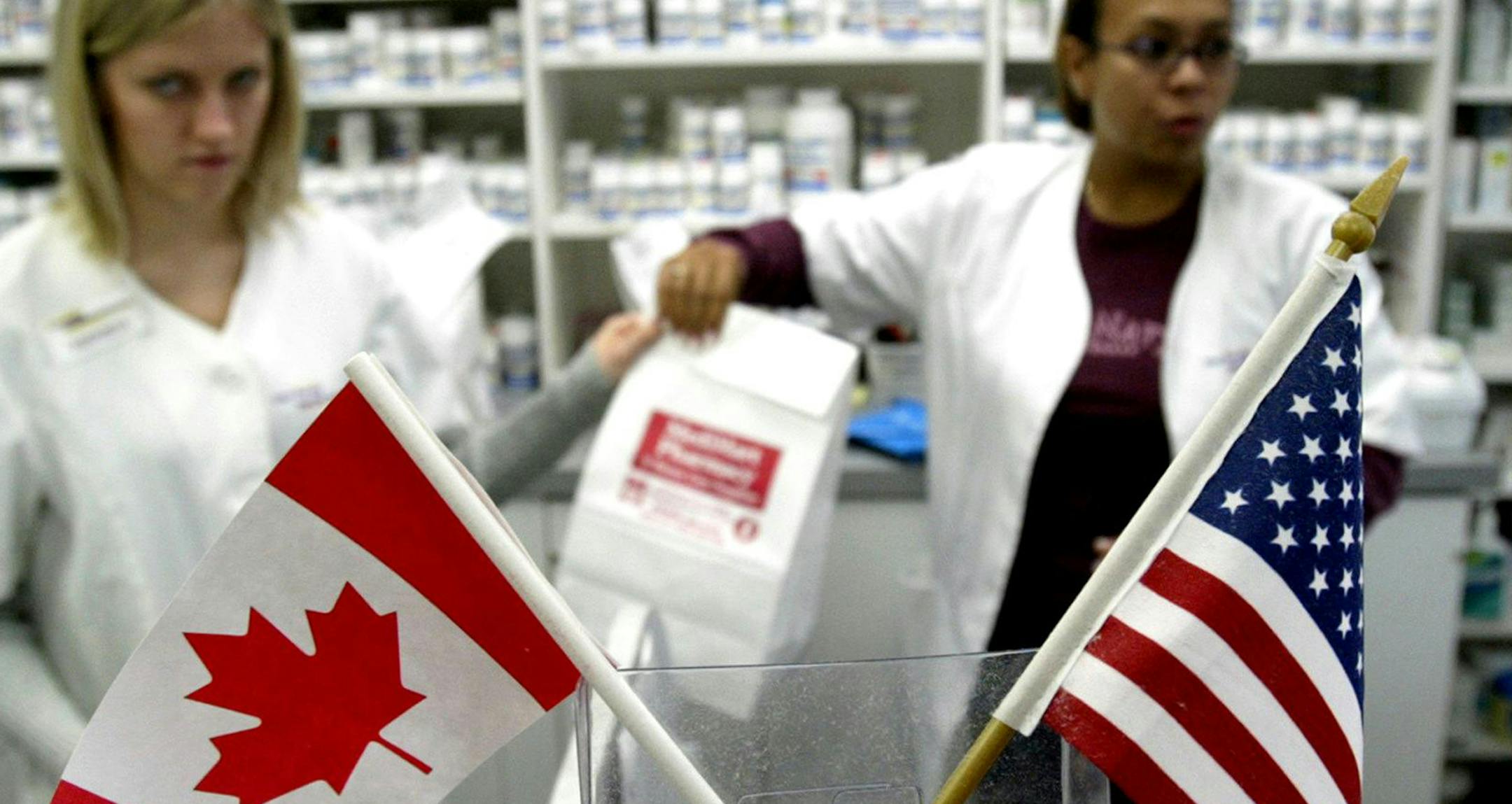 In this file image, Canadian and American flags sit in the foreground as Canadian pharmacist Amanda Hutchinson, left, hands fellow pharmacist Florence Ticne prescription drugs for a group of Minnesota Senior Citizens from the United States on Oct. 22, 2003 who have traveled nine hours by bus across the US-Canadian border to Winnipeg, Canada to buy cheaper drugs. (Jeff Haynes/AFP/Getty Images/TNS)