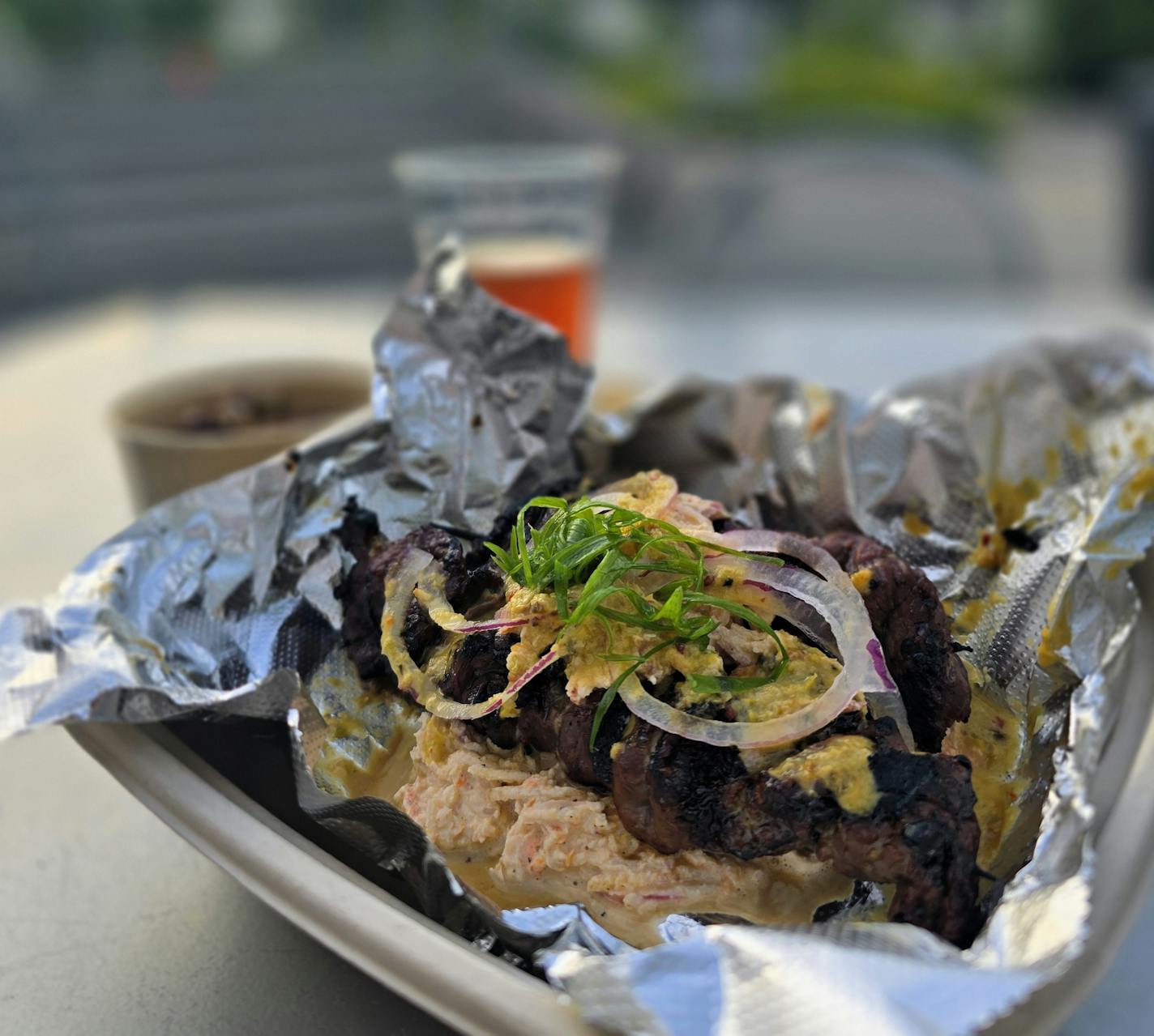 A tray with foil containing two skewers of meet topped with a pink slaw, a beer in the background