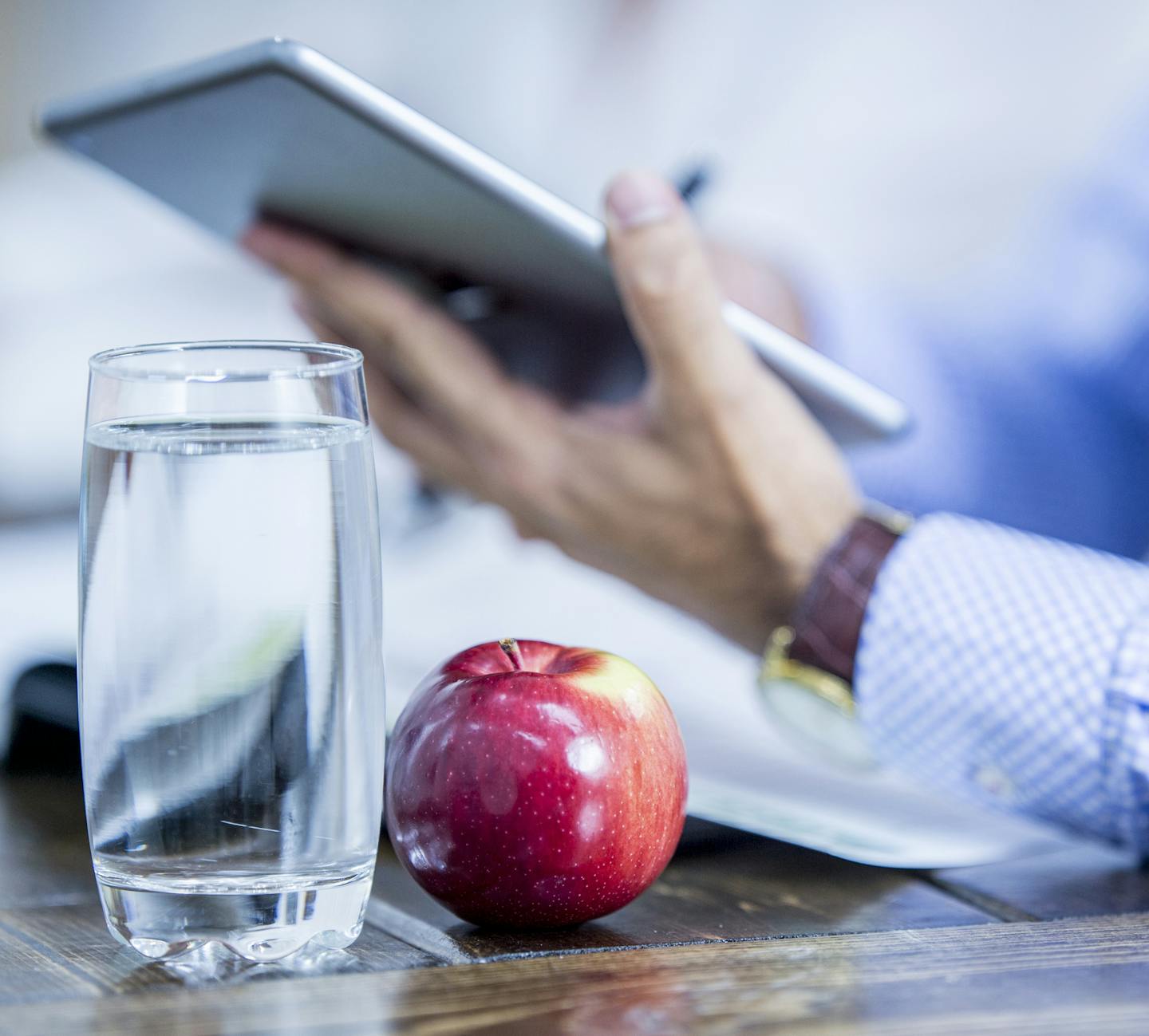 An apple and glass of water are on top of a desk. An office worker is using a tablet computer.