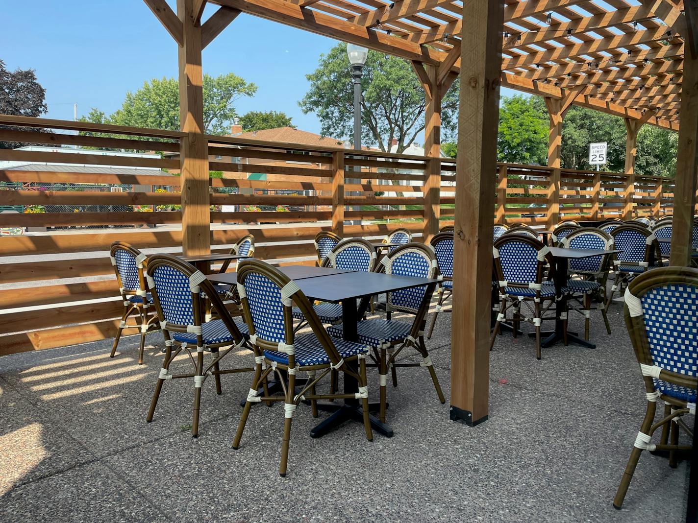 The black with white Parisienne bistro chairs on the sidewalk patio under a wooden cover, surrounded by a fence. Green trees are visible in the background.