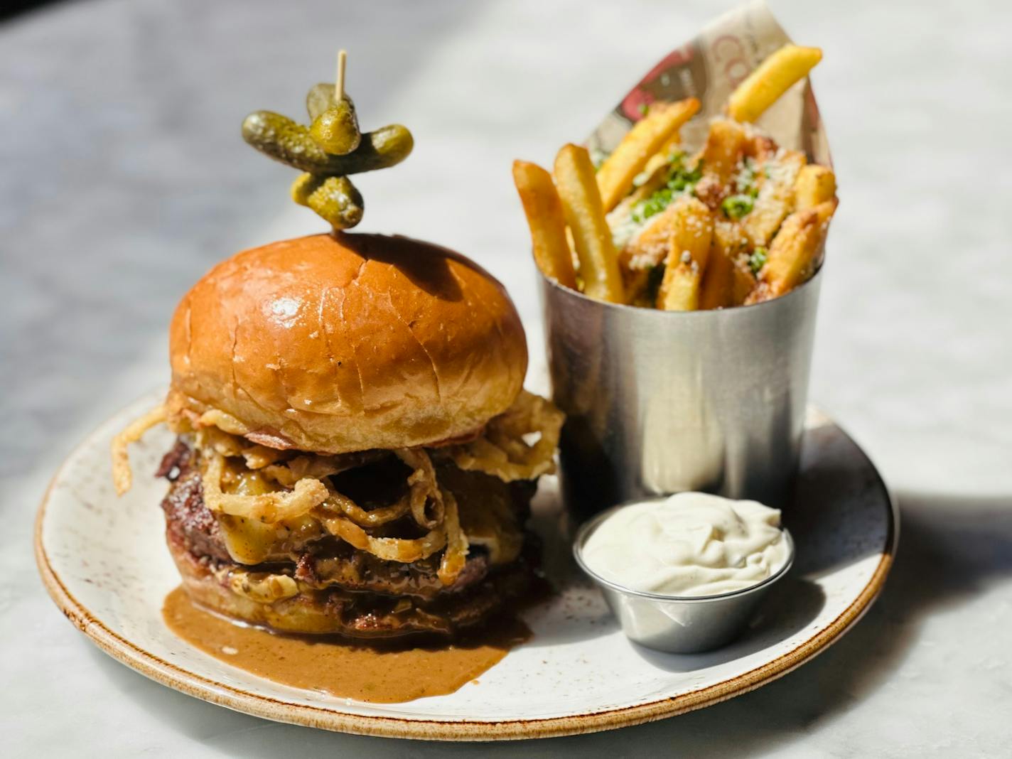 A thick burger on a tall bun is dripping with peppery cream sauce and garnished with a cornichon on a toothpick. Next to the burger is a metal cone of French fries and a small container of truffle-spiked mayo for dipping.