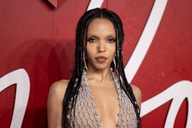 Singer FKA twigs poses against a red backdrop while wearing a silver braided halter gown.