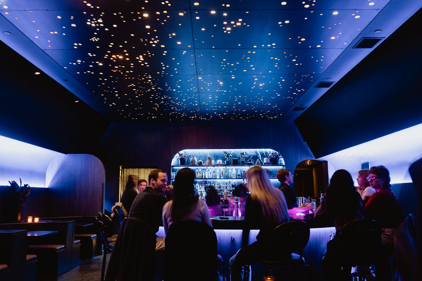 The interior of Stargazer bar with constellation ceiling and a horseshoe shaped bar filled with people in dim purple and blue light.