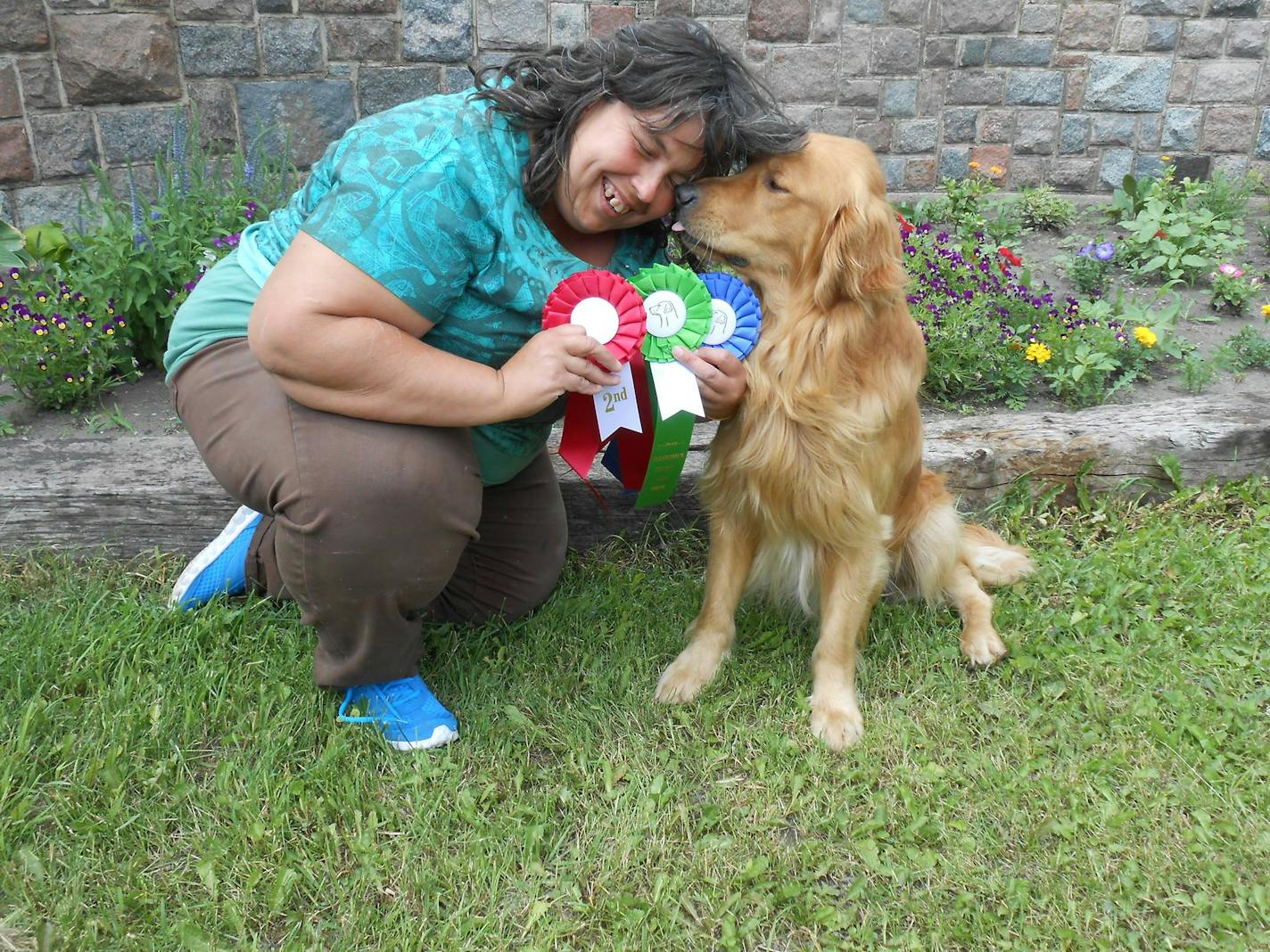 A woman smiles while kneeling down as her dog reaches in to nuzzle her face. She is holding three ribbons in red, green and blue.