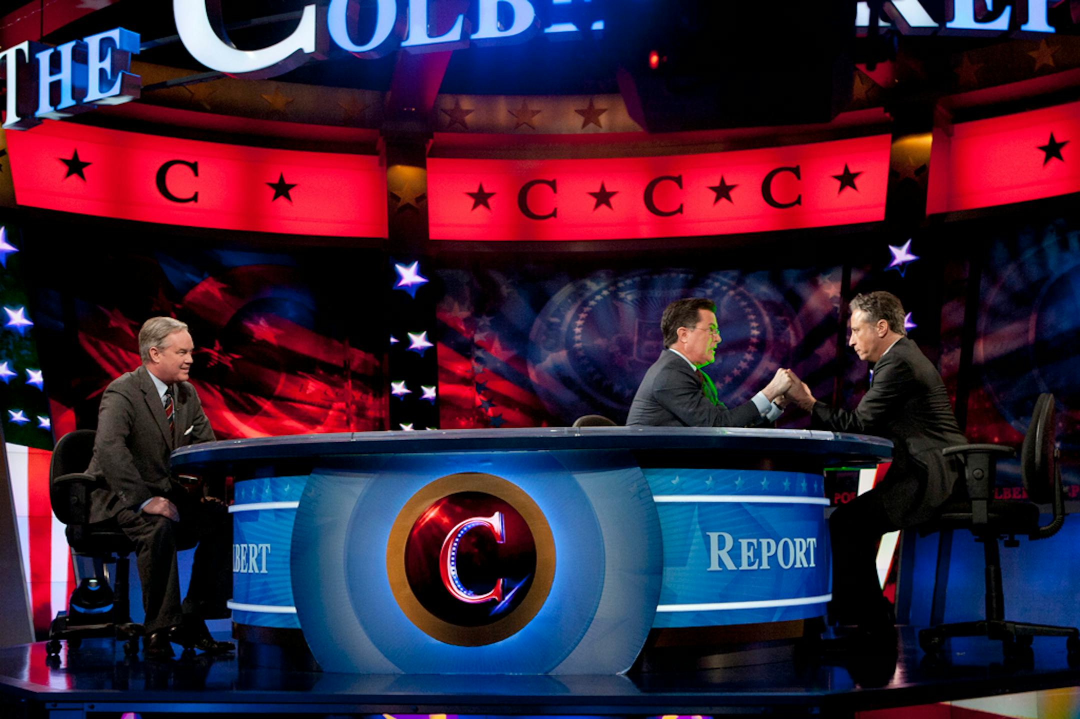 Stephen Colbert, center, and John Stewart, right, hold hands during The Colbert Report, as Trevor Potter looks on Thursday, Jan. 12, 2012, in New York. During the episode, Colbert legally transferred his super political action committee to Stewart, his friend and Comedy Central cohort.