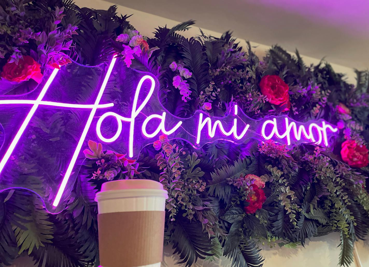 A to-go cup of coffee is held in front of a neon sign that says, "Hola mi amor."