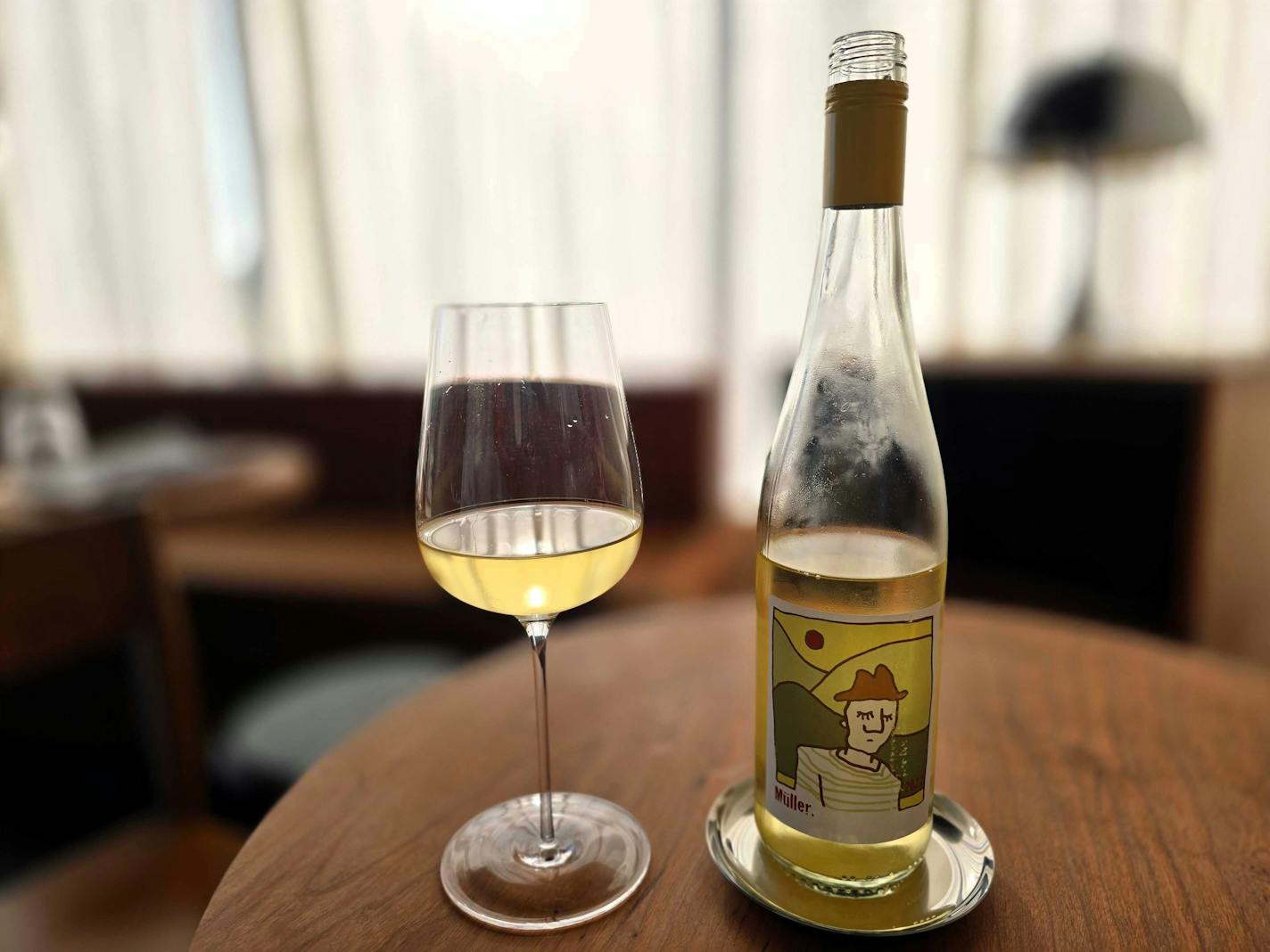 A glass of yellow-hued white wine on a wood table next to a half-poured bottle, in front of a white-curtained window