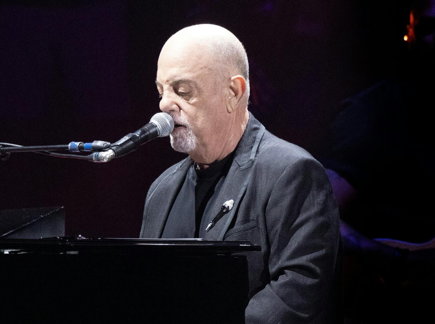 Billy Joel, in gray suit and dark shirt, plays piano and sings into a microphone on a darkened stage.