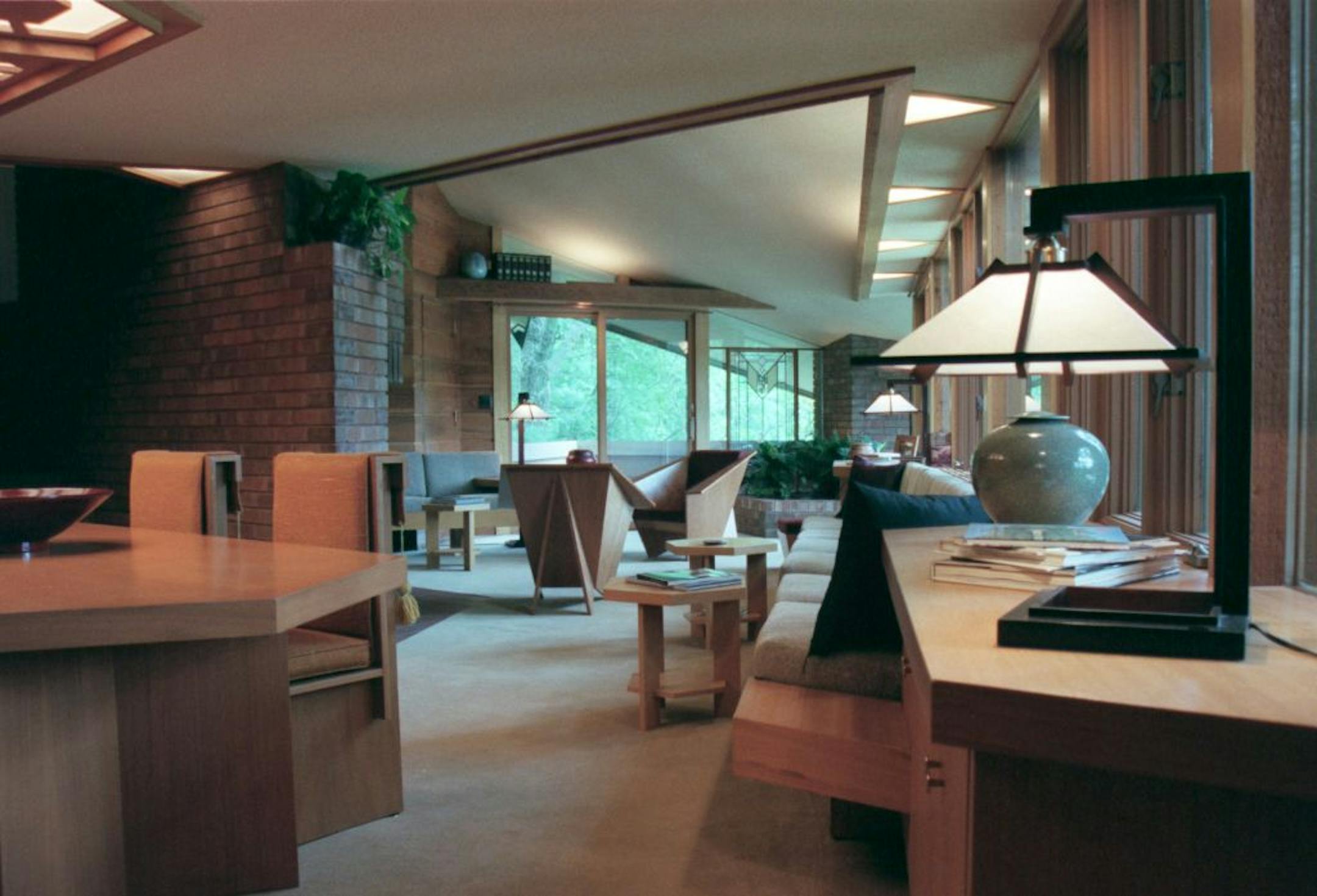 Interior view of the main floor living room area, the dining room table and light fixture to the left side, windows with a view of the pond along the right side, and two � oragami� chairs in front of the brick fireplace ( the chairs are a FLW project to design a chair from a piuece of plywood, ).