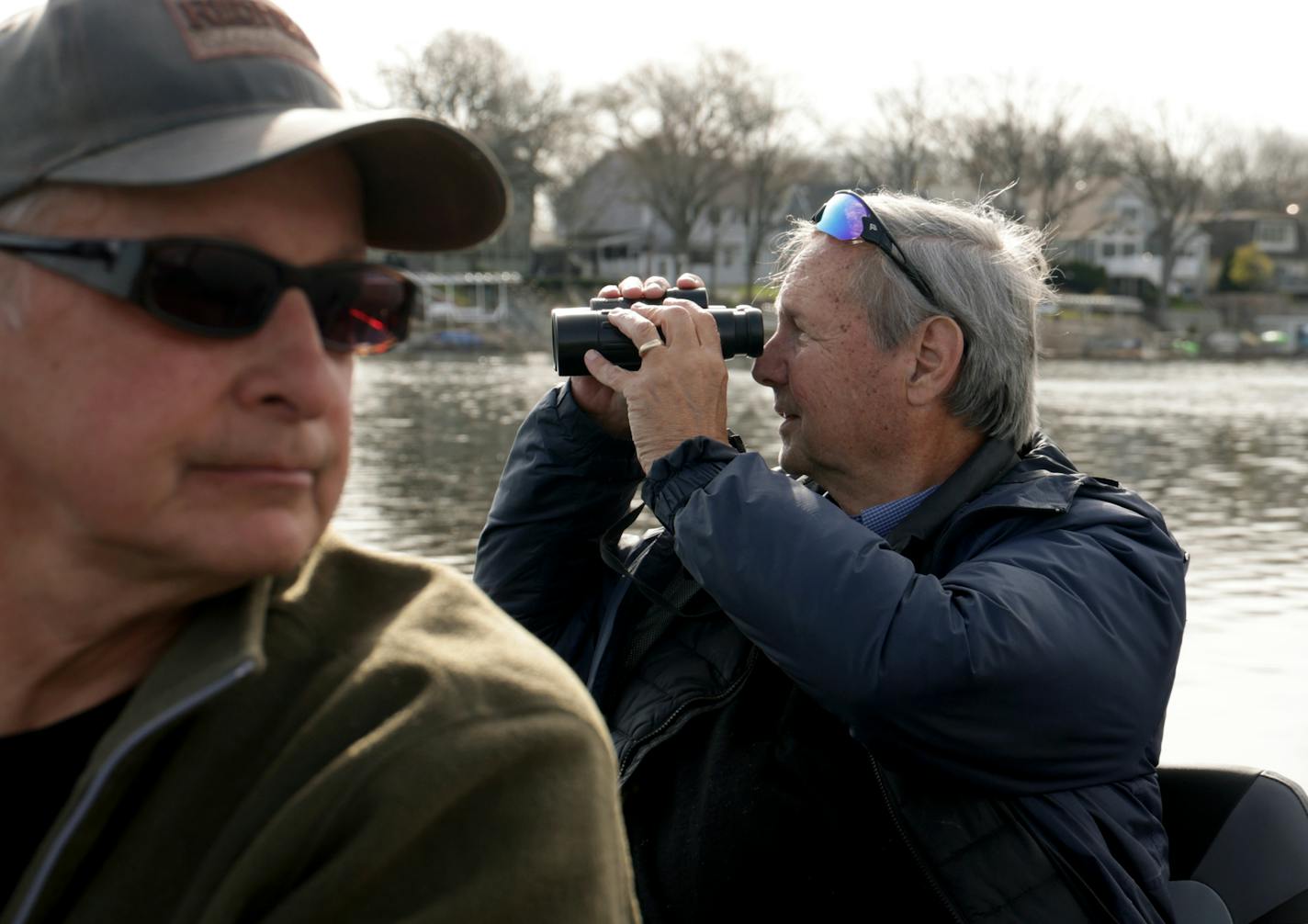 Two men in a boat, one holding binoculars to his face, looking for loons on a lake.