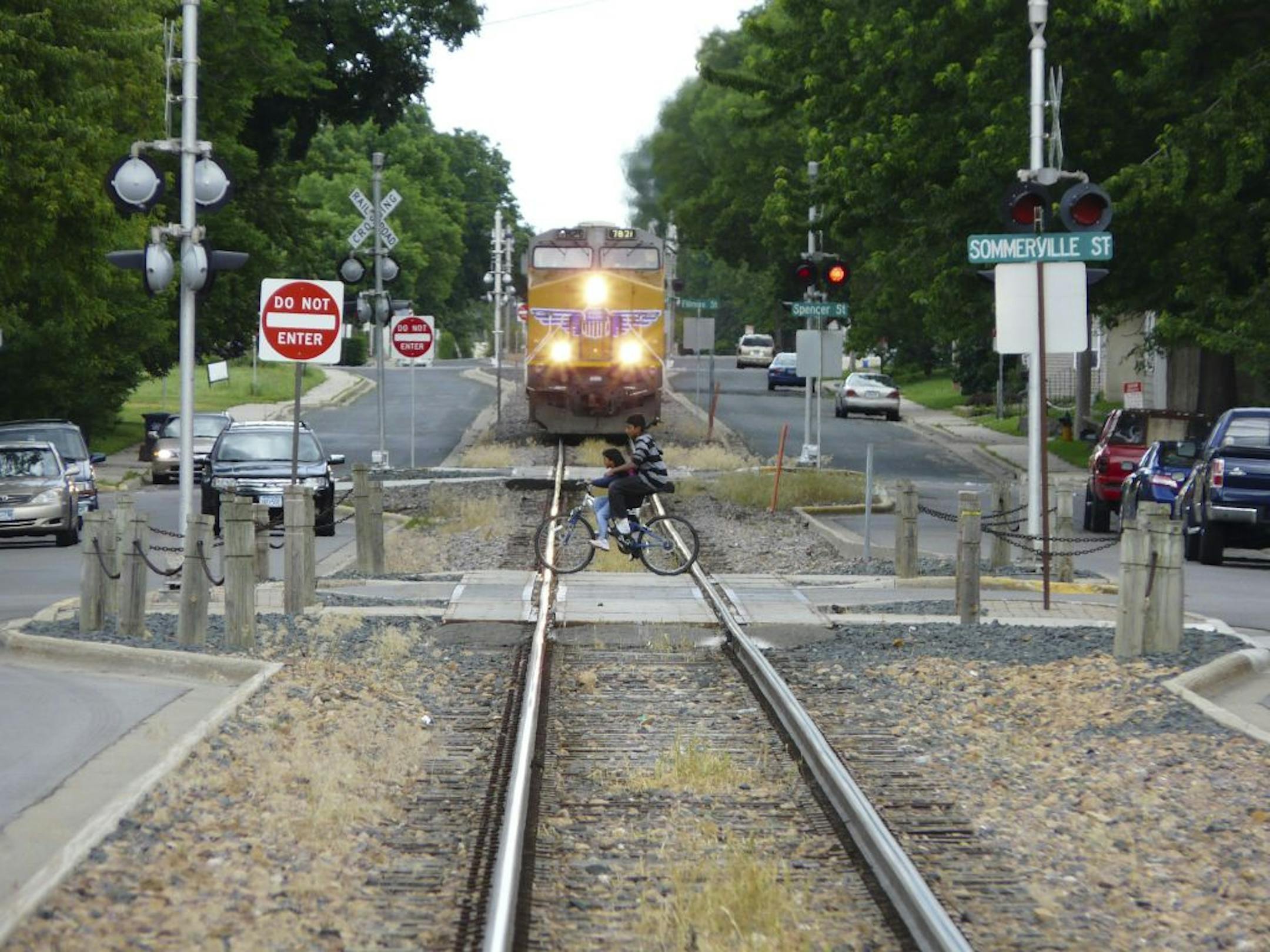 As trains approach in Shakopee when school is out, kids zip across in time; but the train moves extremely slowly and the telephoto lens here flattens the distance between them.