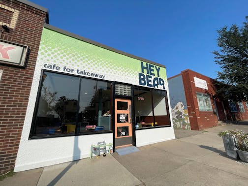 The small outside of the cafe is painted white with a lime green top. There are large windows facing the street and a small, wood framed door.