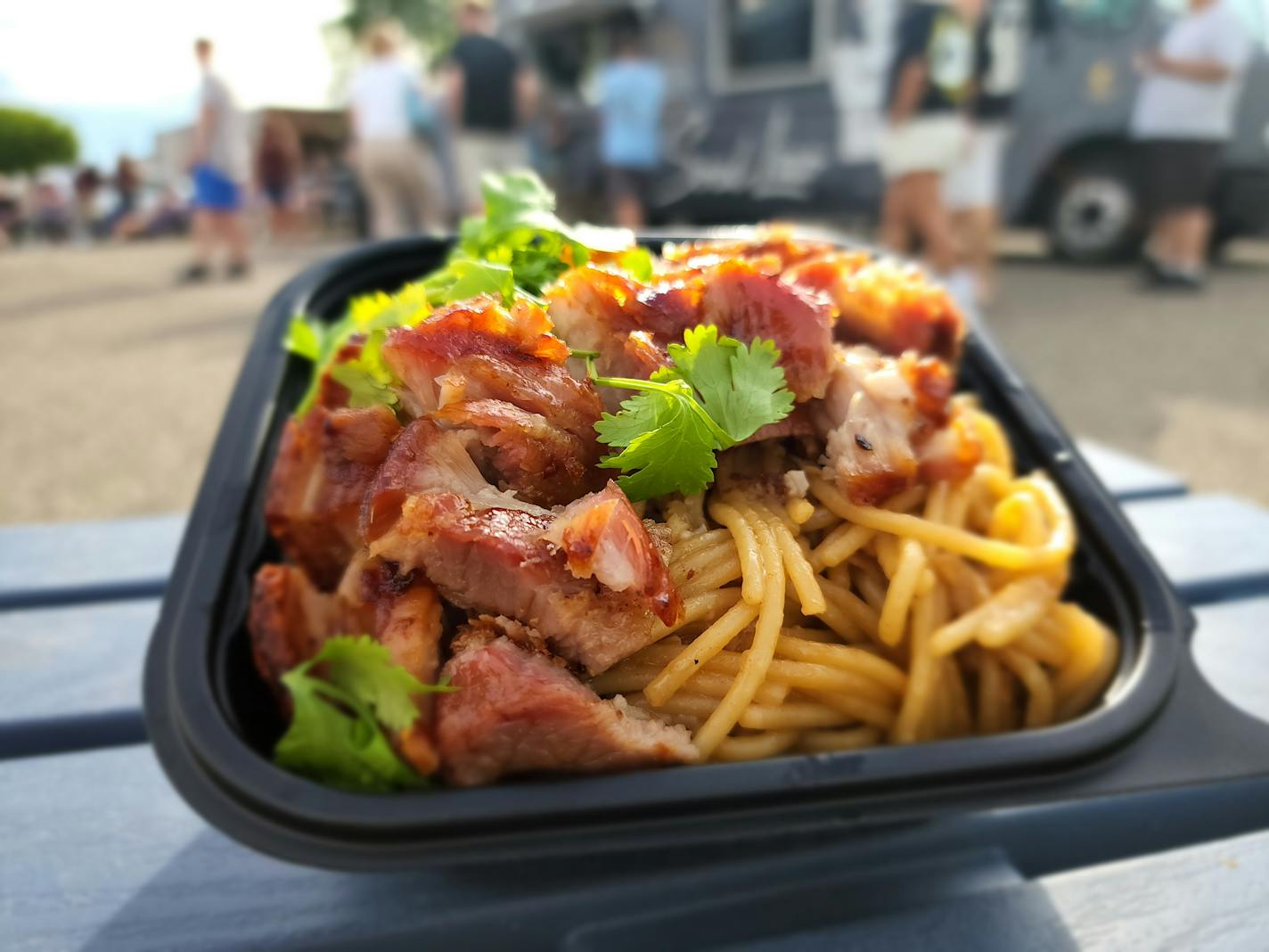 A takeout container of noodles, crispy pork belly and cilantro leaves on a table, with a food truck in the background