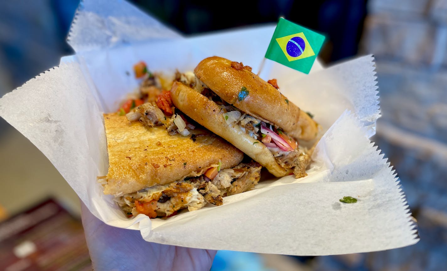 Two slices of a sandwich on a smashed/toasted hoagie roll with bits of pork, tomato, onions, herbs and cheese spilling out the sides. At the tip is a little. Brazilian flag on a toothpick.