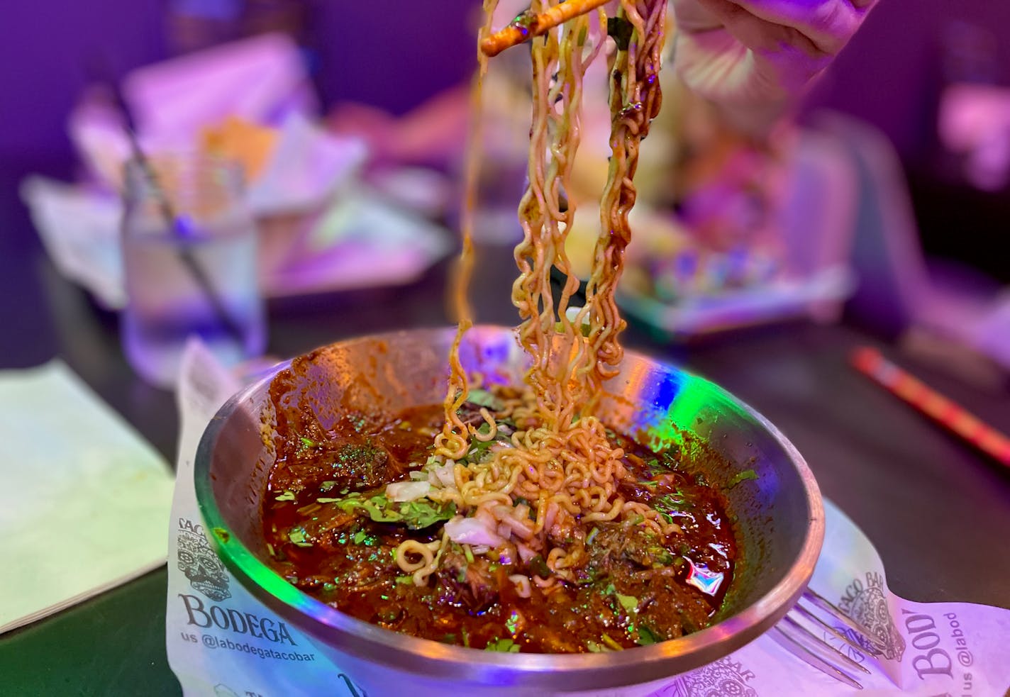 A stainless steel bowl of rich red broth and swiggly ramen noodles flecked with cilantro in the foreground. A hand draws up a tangle of noodles with saucy chopsticks.