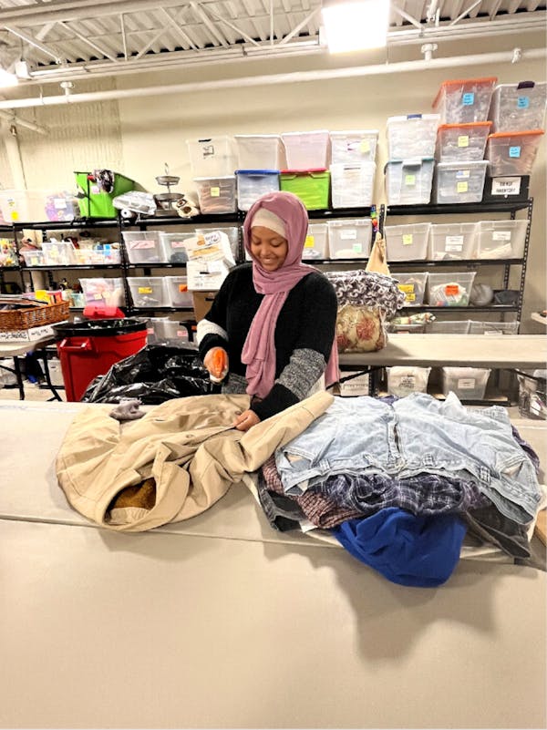 A young woman stands at a table in a storage room. She's pointing a spray bottle at a coat laid out on the table alongside a pile of other clothing items.