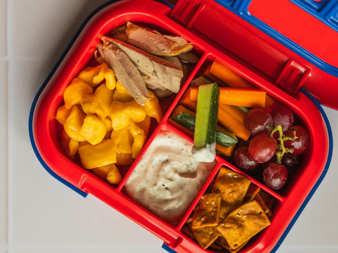 A bright red and blue bento box lunch container with smoked meat, cheese, vegetables, crackers and dip.