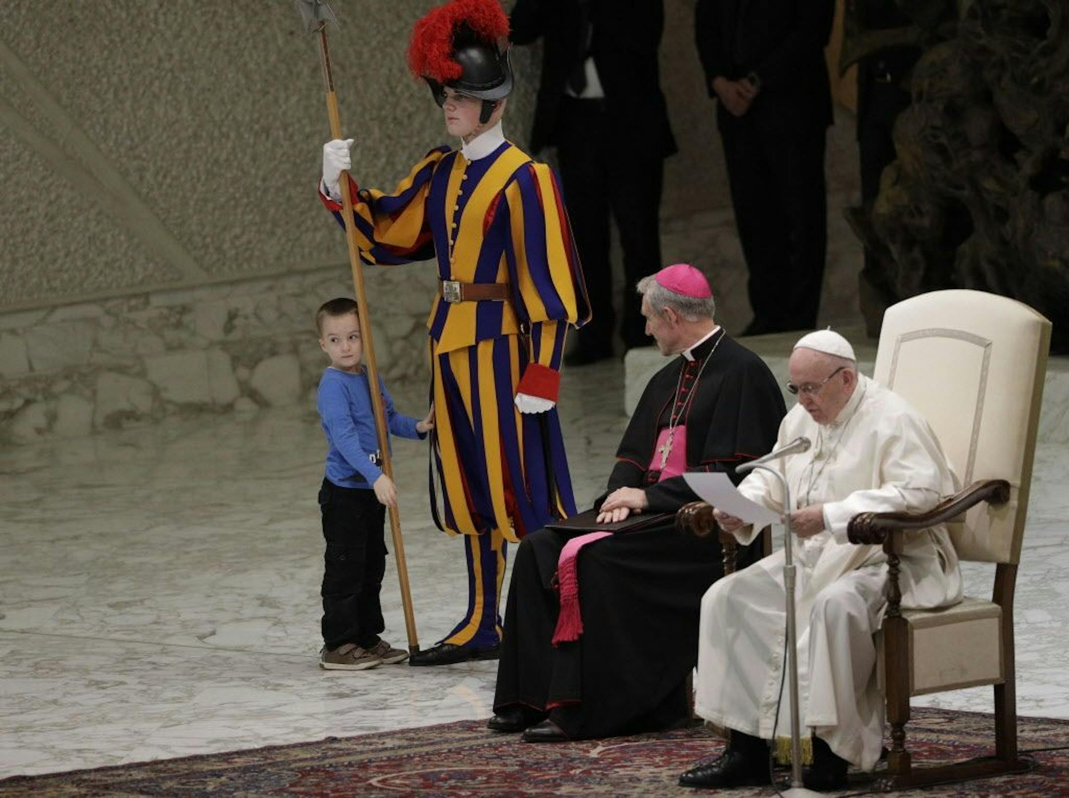 A child stands by a Swiss guard after getting up to the area where Pope Francis and Archbishop George Gaenswein are sitting, during the pontiff' weekly general audience in the Paul VI Hall at the Vatican, Wednesday, Nov. 28, 2018. Pope Francis has praised the freedom, albeit undisciplined, of a hearing impaired child who climbed onto the stage during his general audience to play. The Swiss Guards and Vatican gendarmes stood by Wednesday and gamely let the young boy run around Francis as monsigno