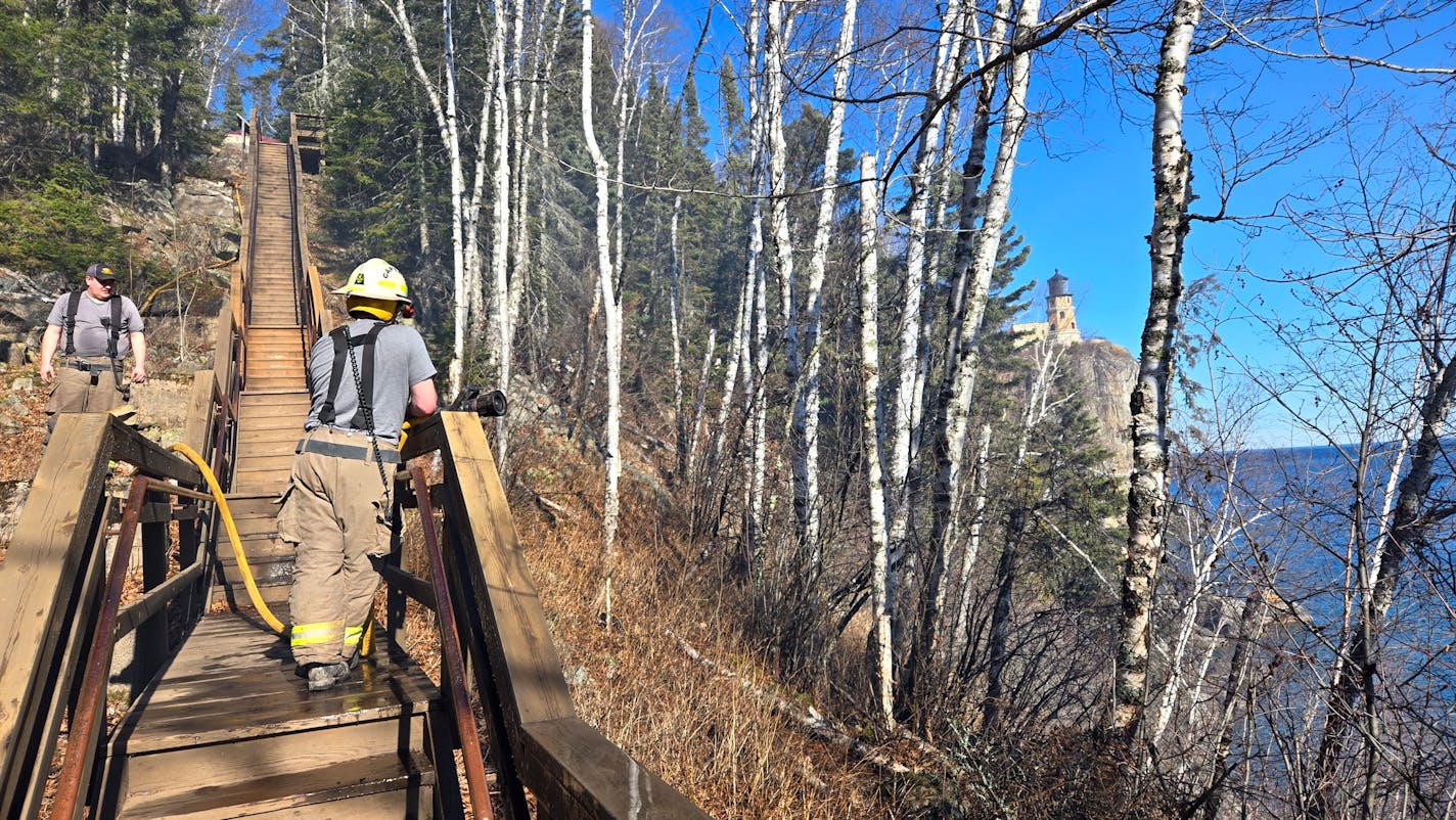 firefighters stand on a wooden stairwell. to their right, a lighthouse sits atop an outcropping of rock