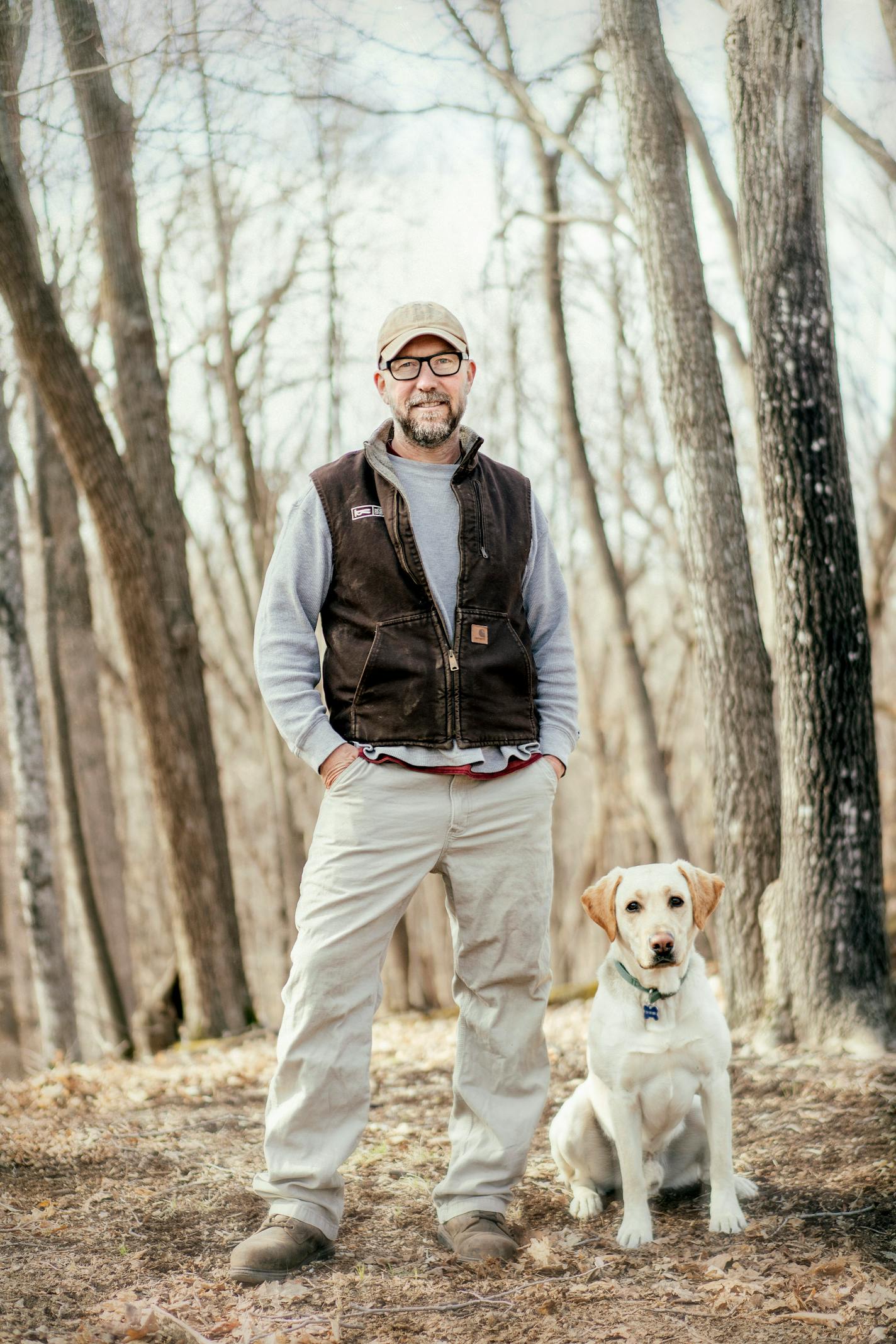 A man wearing a ball cap, dark-rimmed glasses, vest and work pants stands in the woods besides a yellow Lab.