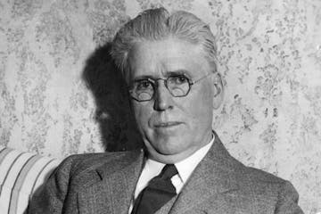 Former St. Paul Mayor William Mahoney wearing a suit and tie and seated on striped sofa in front of wall with wallpaper.