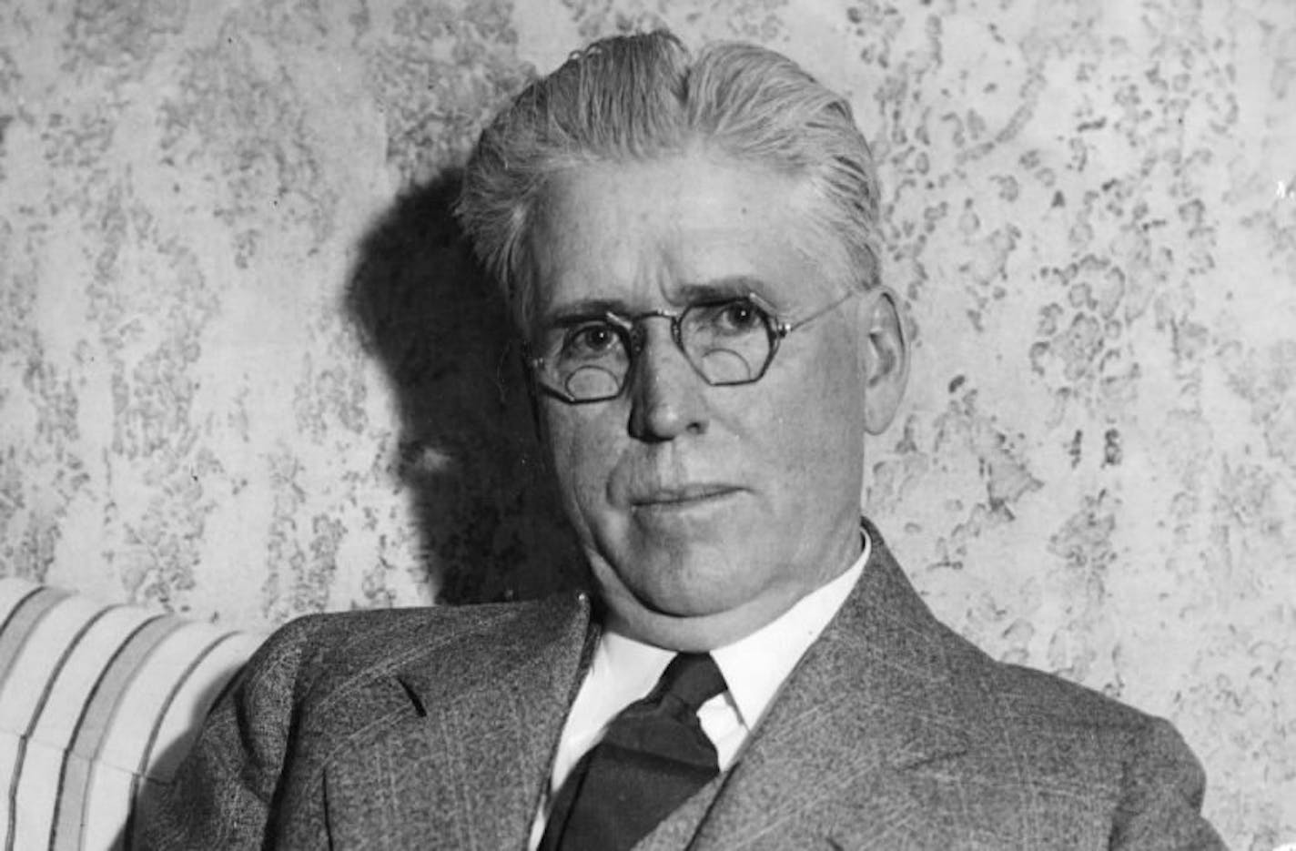 Former St. Paul Mayor William Mahoney wearing a suit and tie and seated on striped sofa in front of wall with wallpaper.