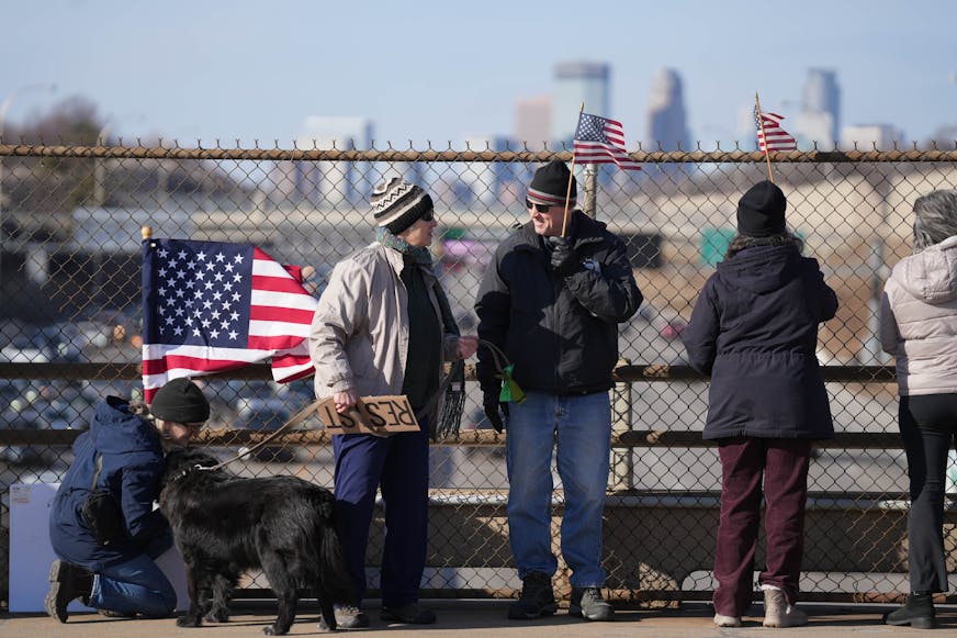 A group of people standing on a pedestrian bridge over a highway are waving American flags. One woman is holding a protest sign saying "RESIST."