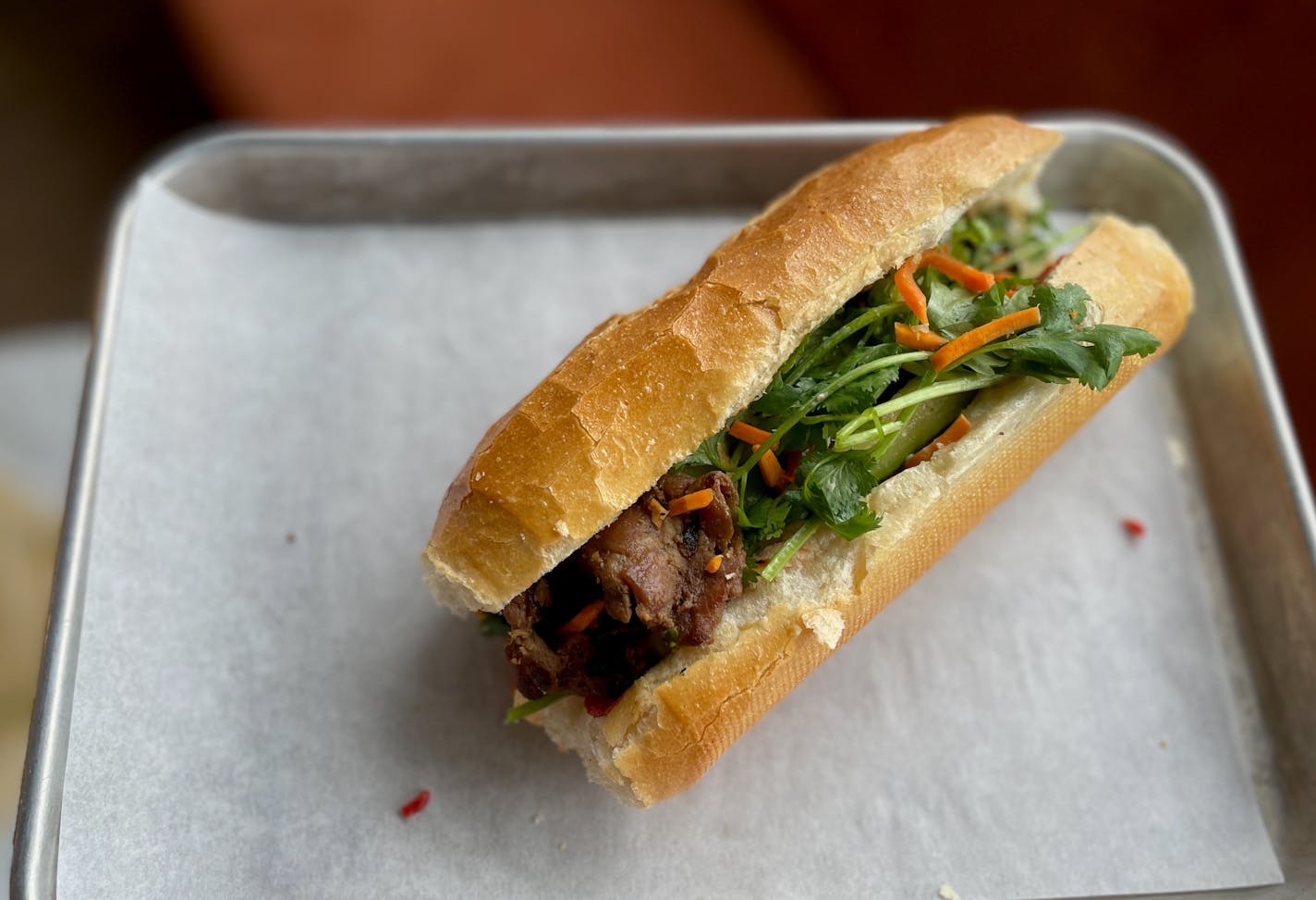 On a baking tray lined with plain wax paper is a sandwich on a crackled, toasty looking baguette with pickled carrots, cilantro and roasted meat peeking out.