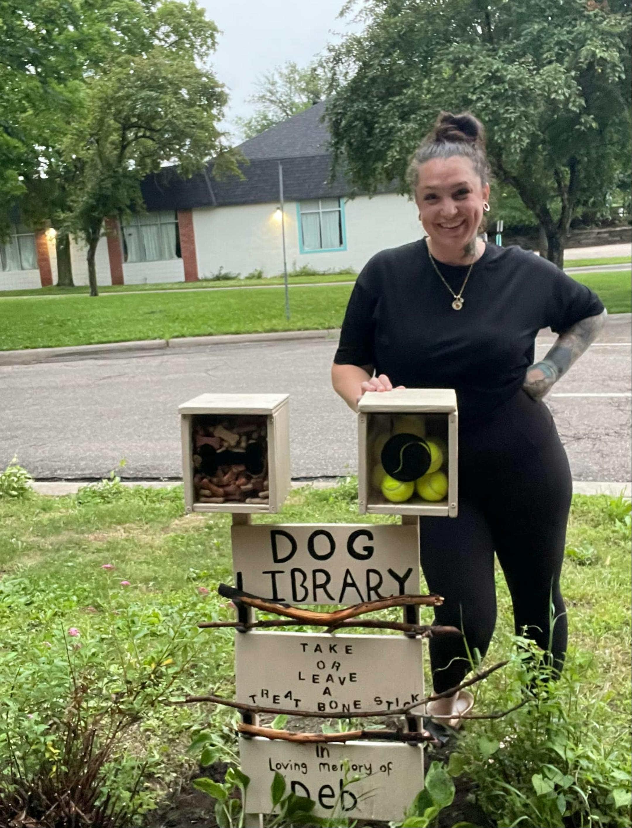 Woman in black T-shirt and leggings smiles for camera behind a stand supporting a box of tennis balls, a box of dog treats, and shelves for sticks.