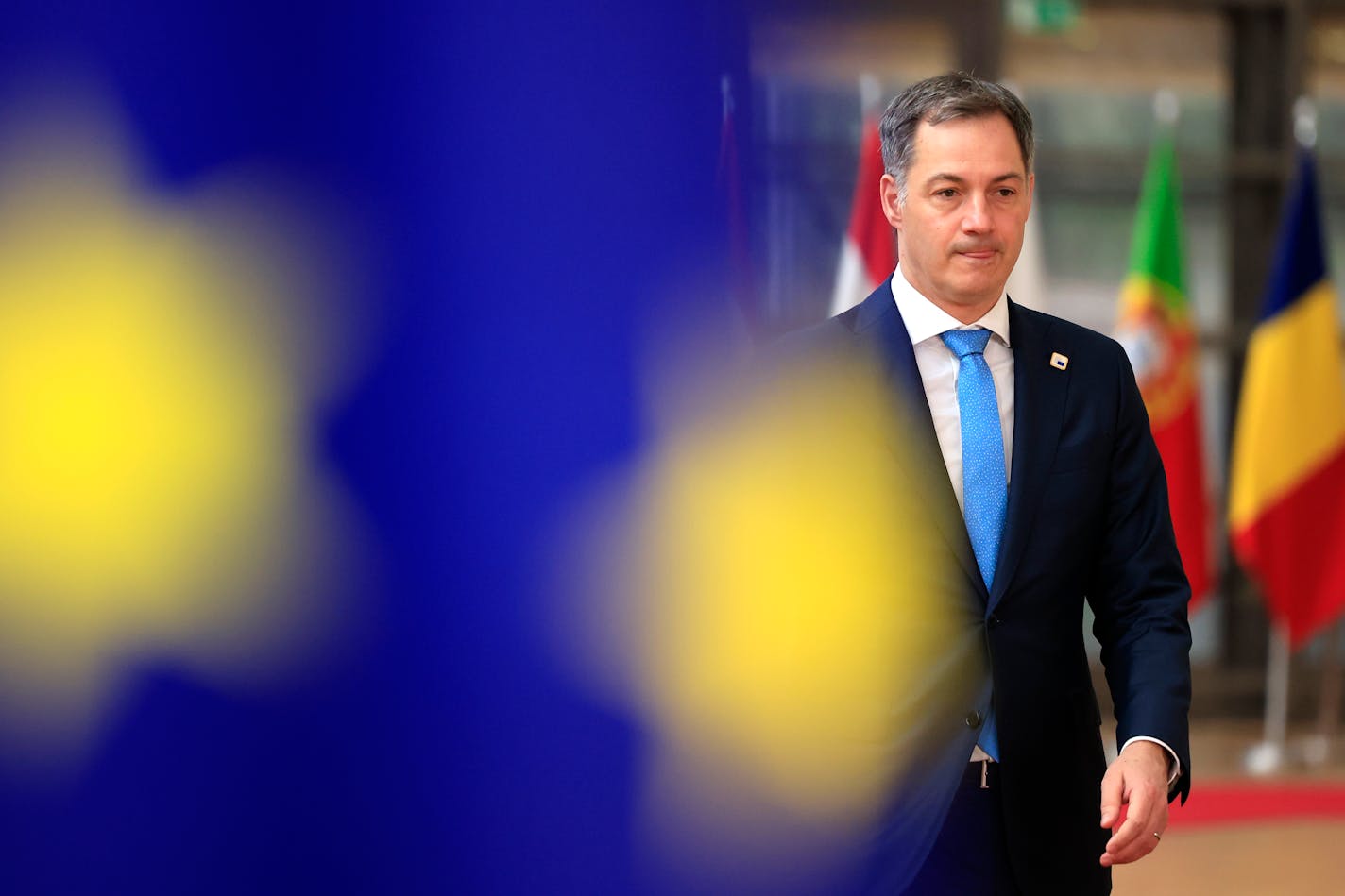 Belgium's Prime Minister Alexander De Croo in navy suit with white shirt and sky blue tie with pin on lapel with blurred blue and yellow flag in foreground and other flags in background.