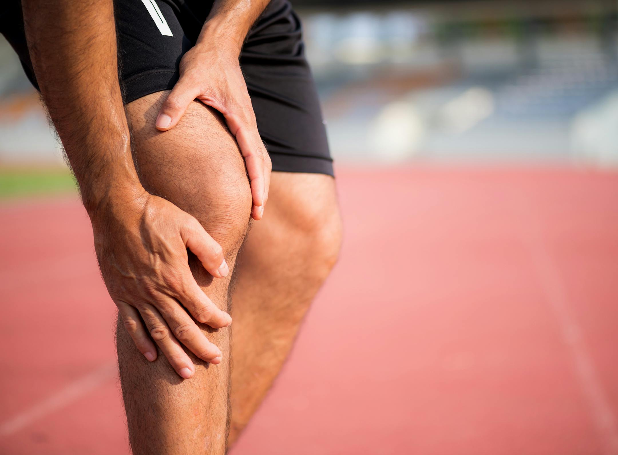 knee Injuries. young sport man with strong athletic legs holding knee with his hands in pain after suffering muscle injury during a running workout training on Running Track. Healthcare and sport concept.
