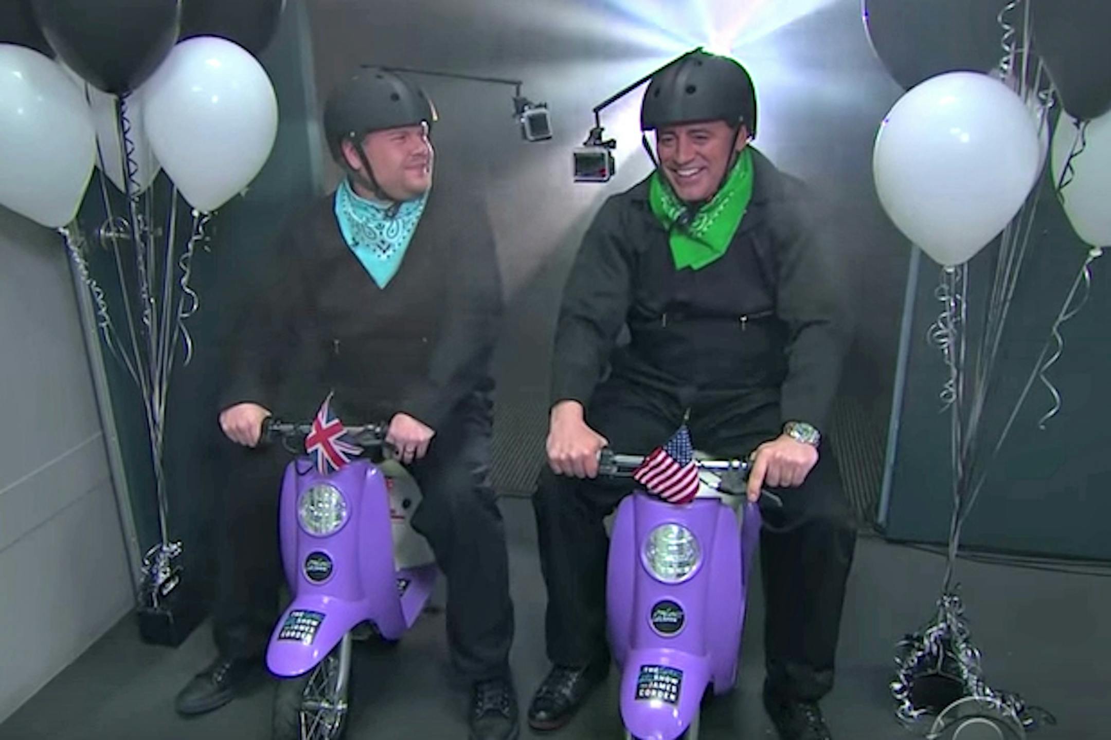 James Corden, left, and Matt LeBlanc race each other on scooters.