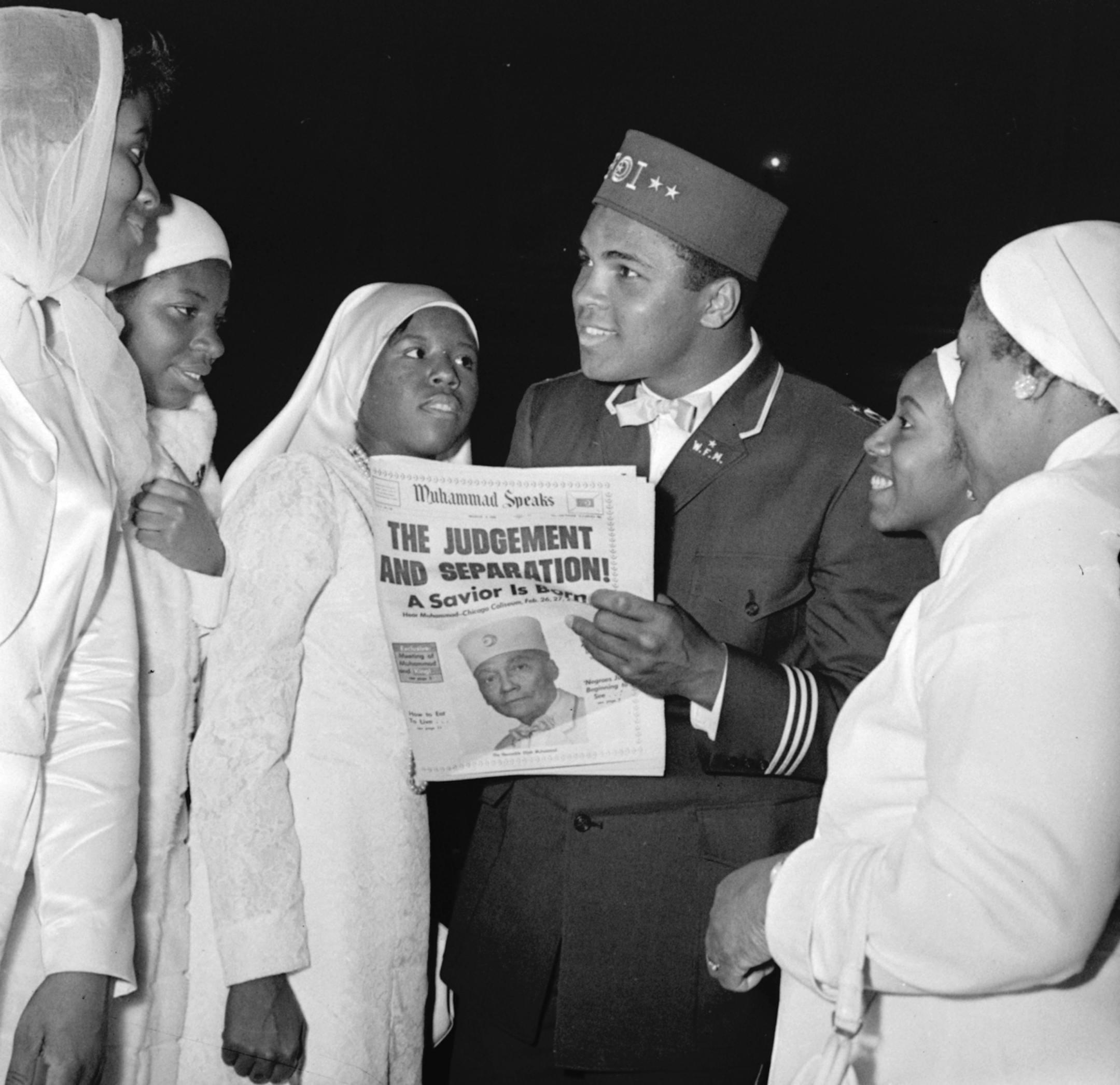Muhammad Ali, world heavyweight champion, wears a uniform of the Black Muslim organization as he attends their convention, February 26, 1966, in Chicago. He is talking with Sisters of Islam, female members of the group who wear the habit of their order. The newspaper held by Ali carries photo of Elijah Muhammad, head of the Black Muslims.