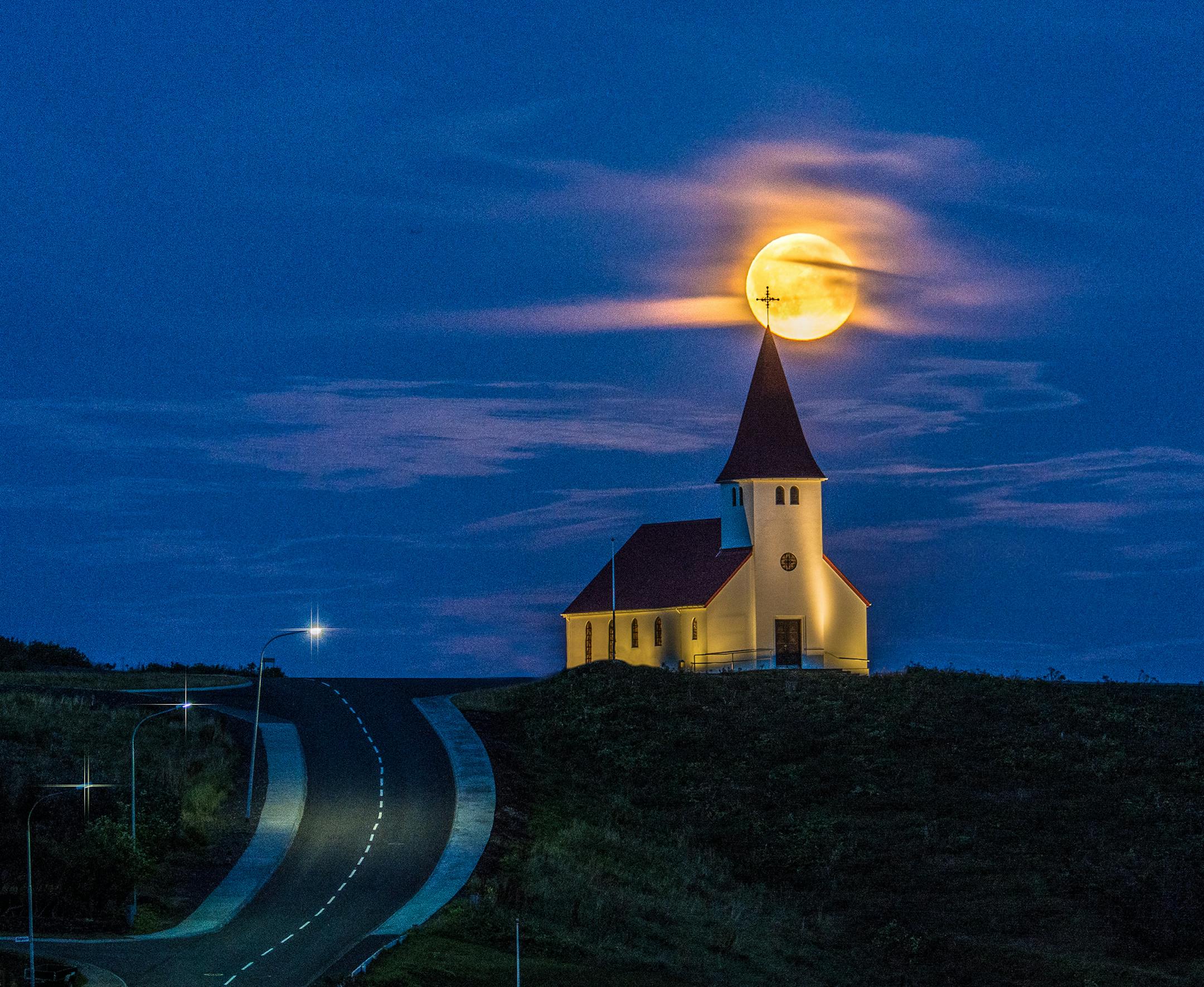 My wife, Beverly and I were vacationing in Iceland. We were headed into Vik for supper when we came around a corner and saw the church with the moon behind it. We quickly stopped the car. I realized I had left my tripod back at our room, so we took turns using Beverlyís. The moon was shifting positions very quickly, so we had to hurry. It was all over in about five minutes. The beauty of the full moon, the ethereal clouds, and the silhouette of the churchís cross appearing against the