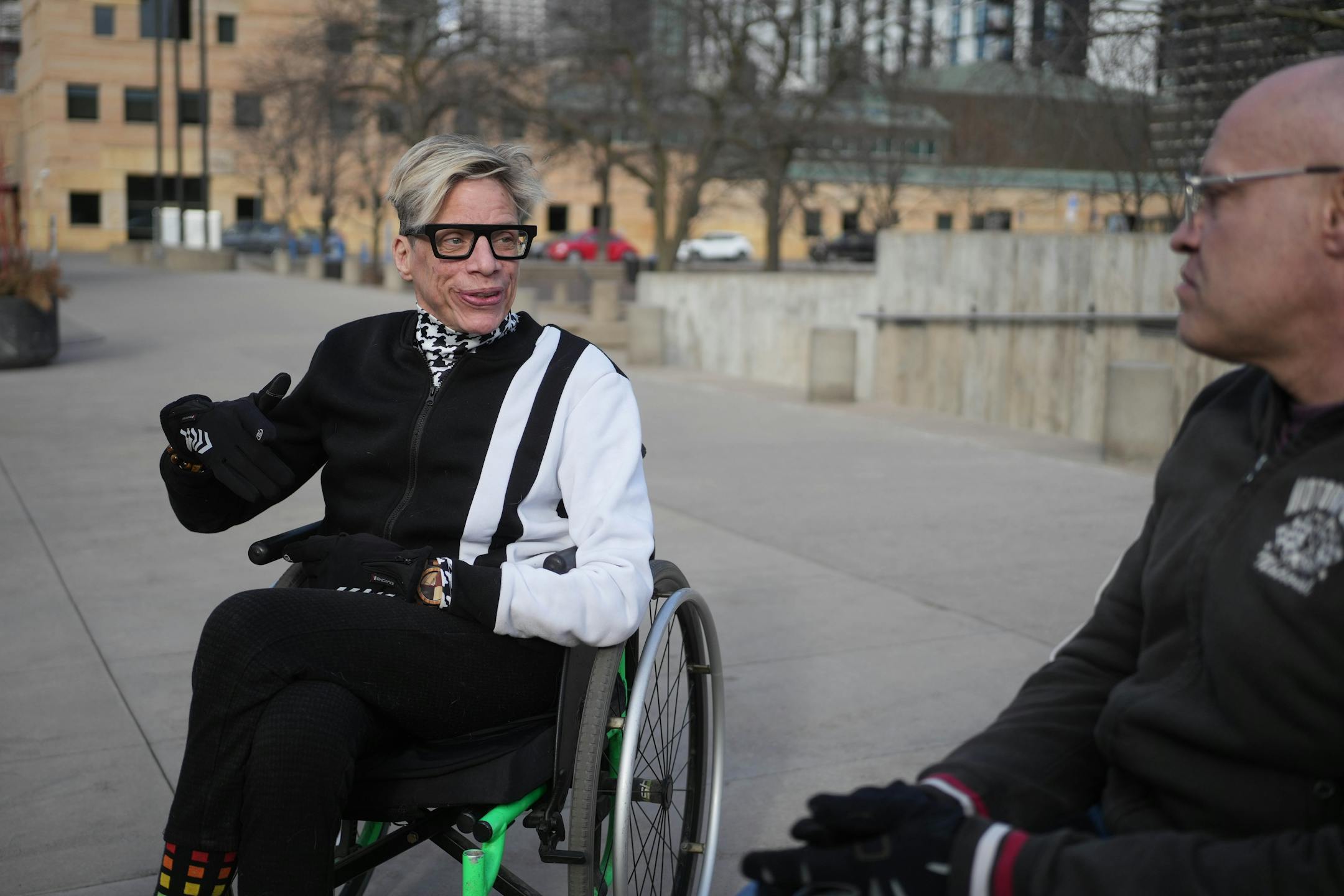 Two men in wheelchairs are talking on a sidewalk.