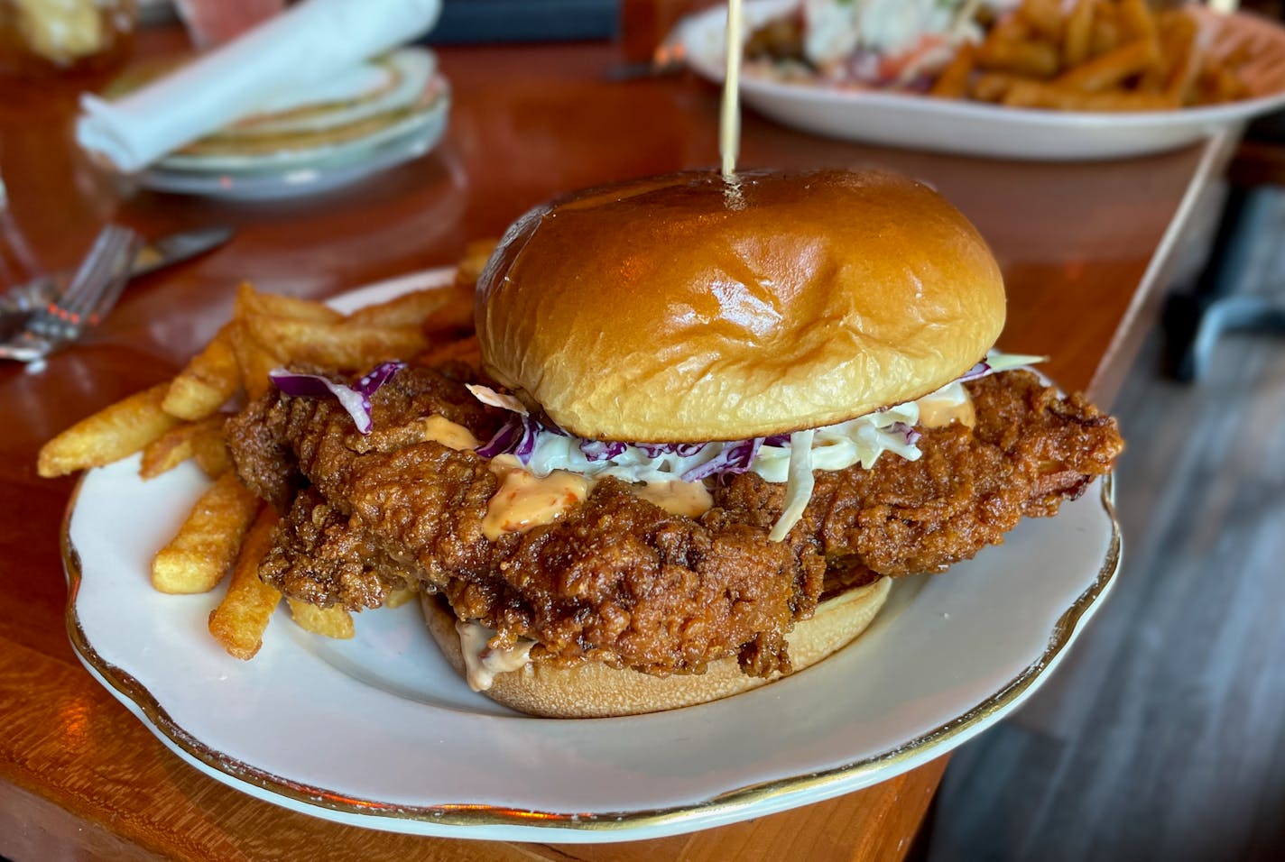 A large fried chicken sandwich dominates a white supper club plate with a side of French fries. Chili-flake spiked mayonnaise is just barely visible with a smattering of red cabbage coleslaw.